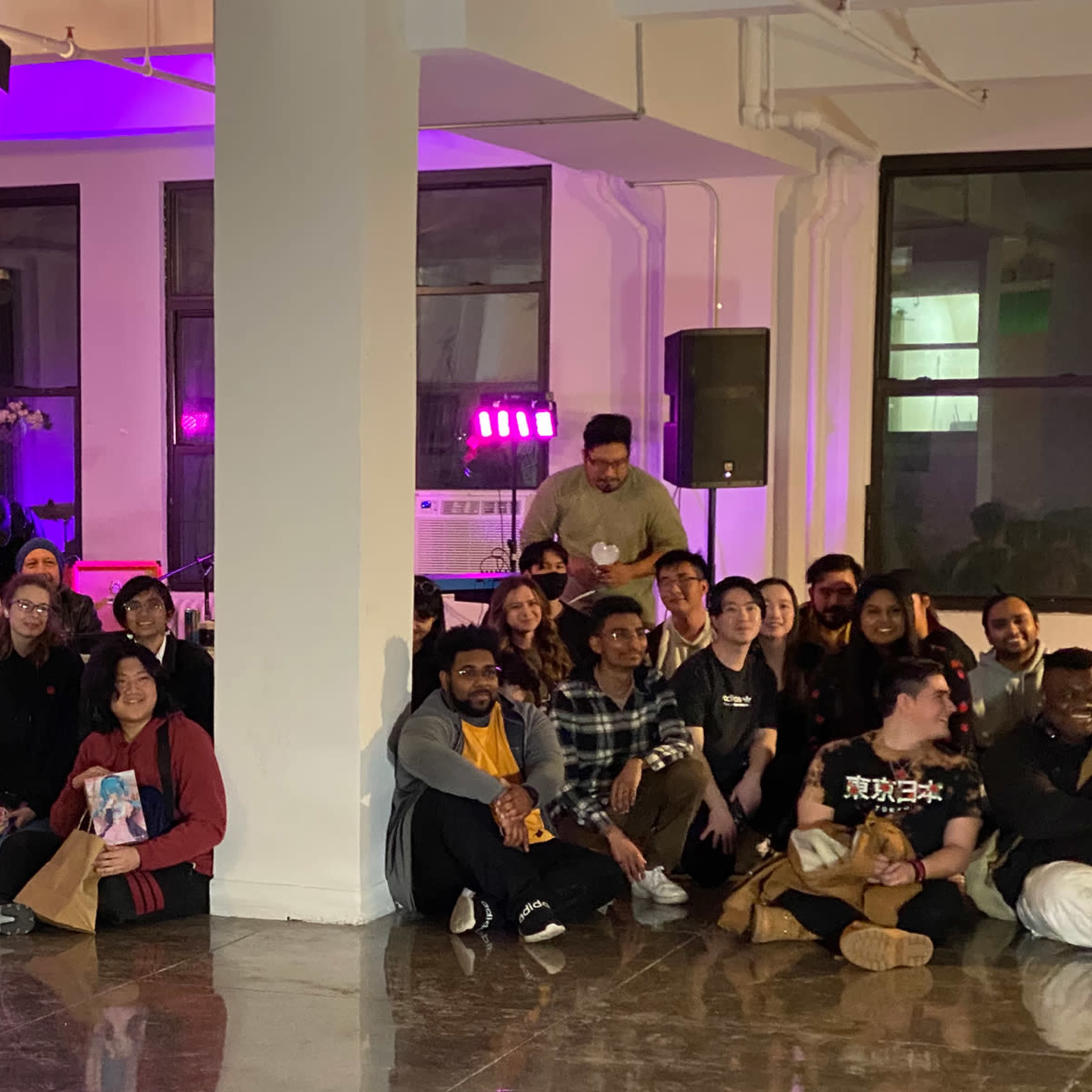 A large group of people poses together in a brightly lit room with a stage set up for a musical performance in the background.