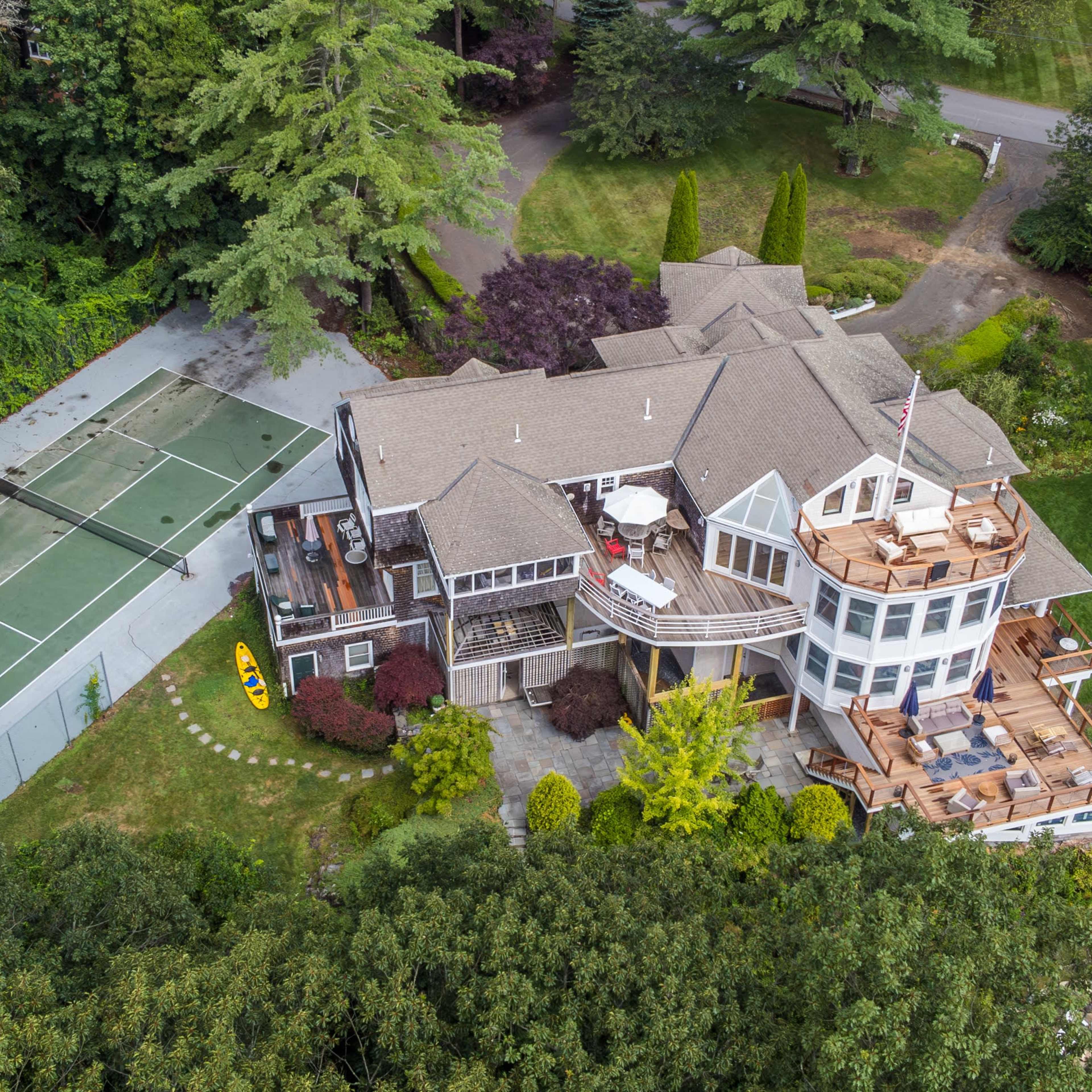 A large, multi-level house with a surrounding green lawn, a wooden deck, and a nearby tennis court.