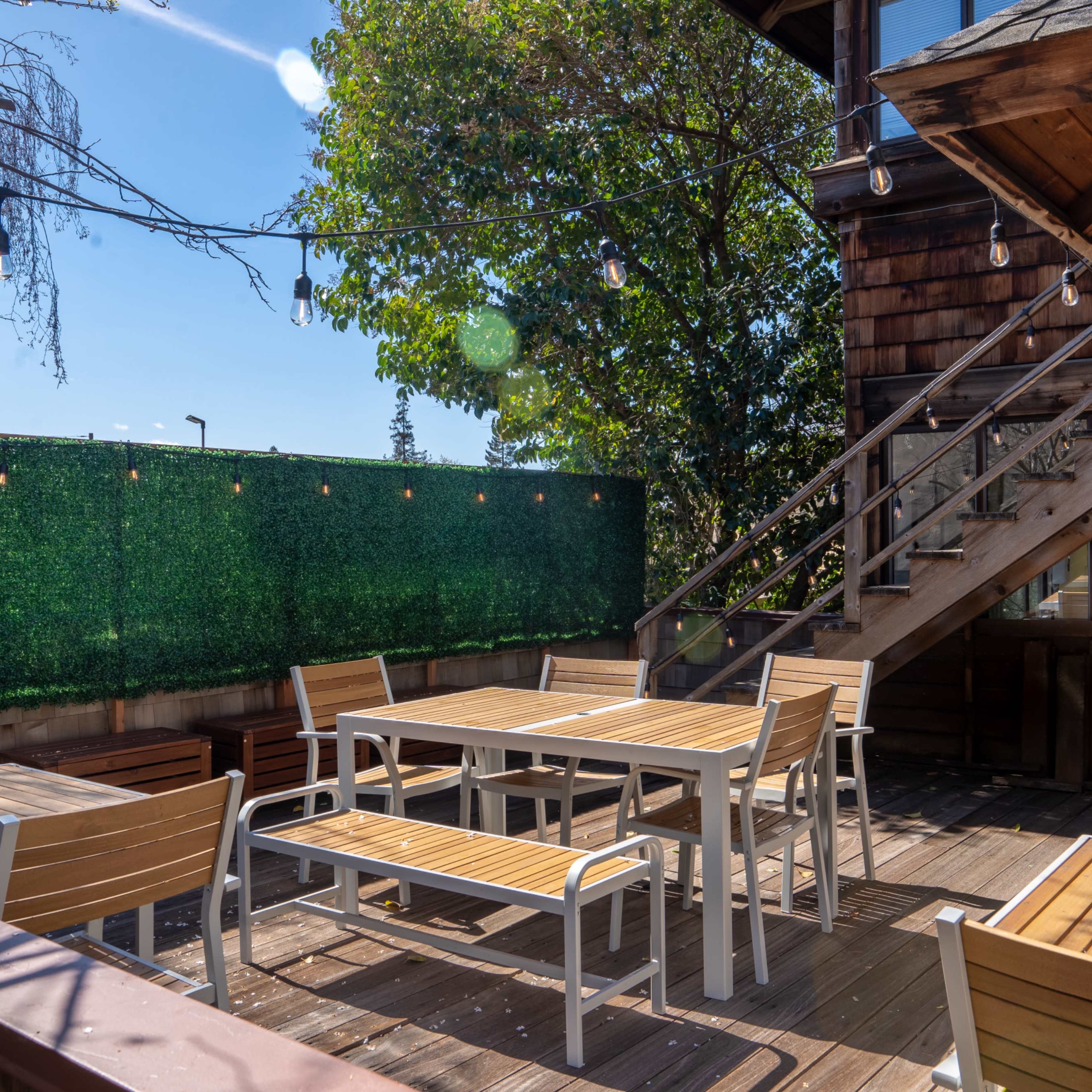 A wooden outdoor patio with several tables and chairs, surrounded by greenery and a staircase leading to an upper level.