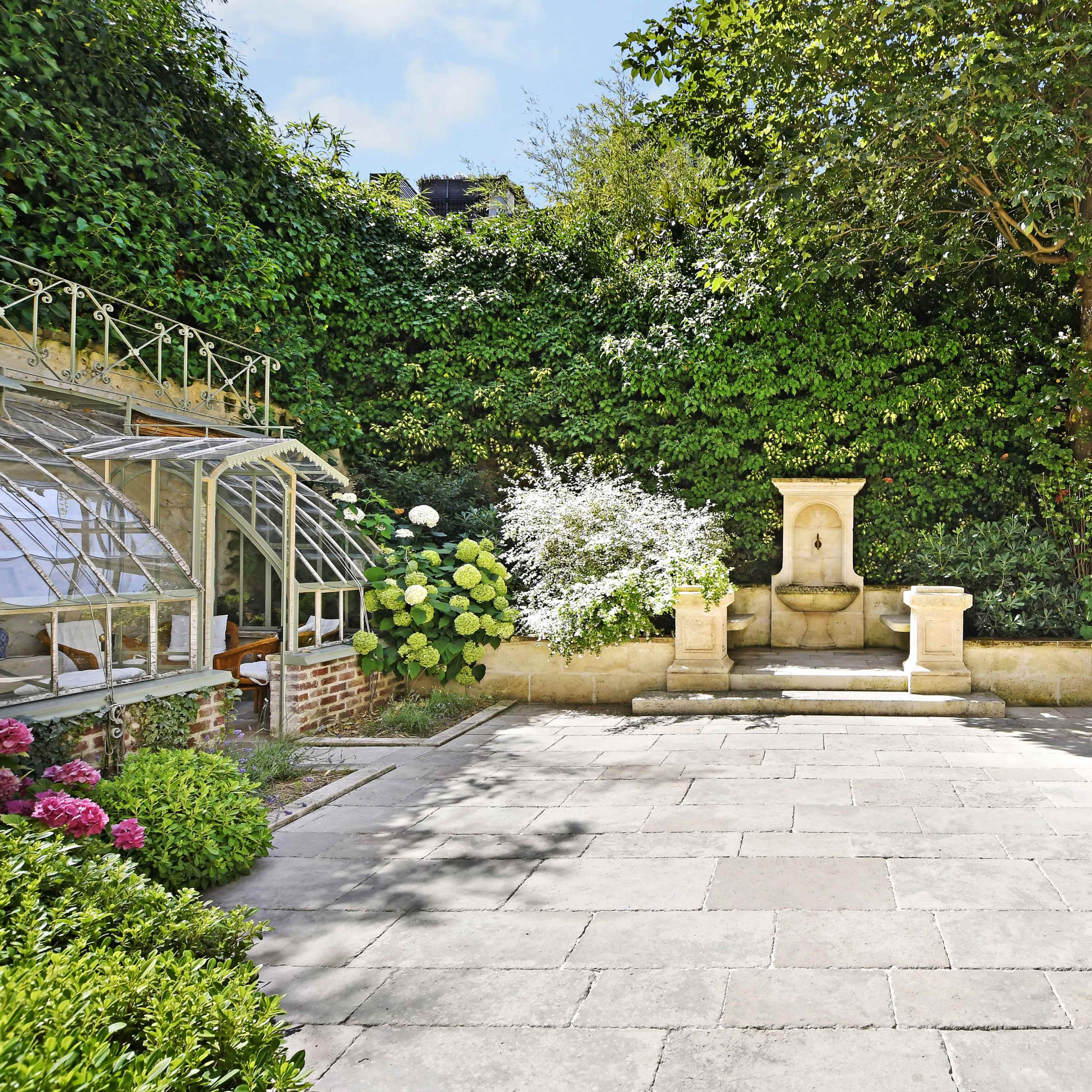 A stone patio surrounded by lush greenery, a fountain, and flowering plants.