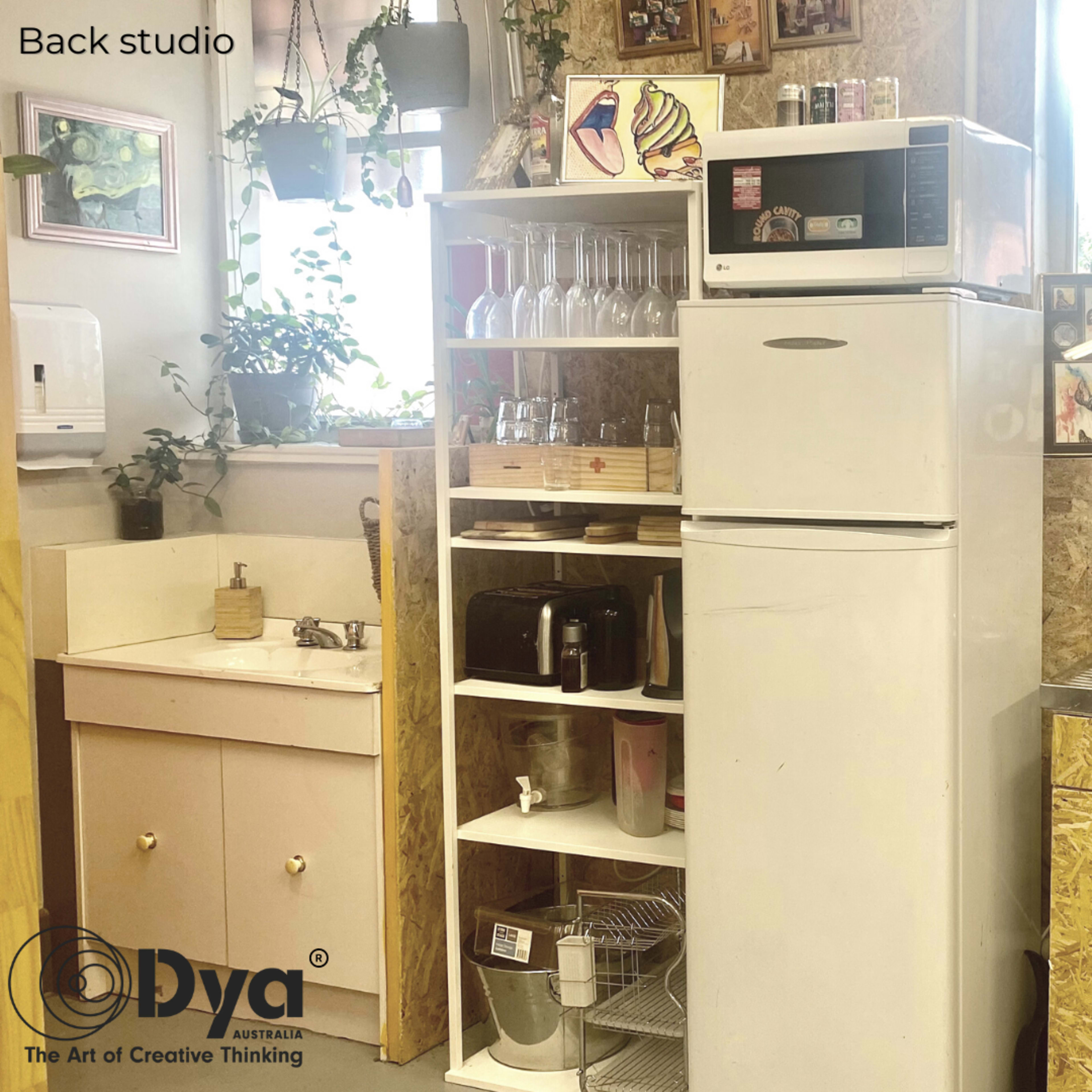 A cluttered kitchen area featuring a white refrigerator, a sink, shelves with glasses and utensils, and a countertop with various appliances in a studio environment.