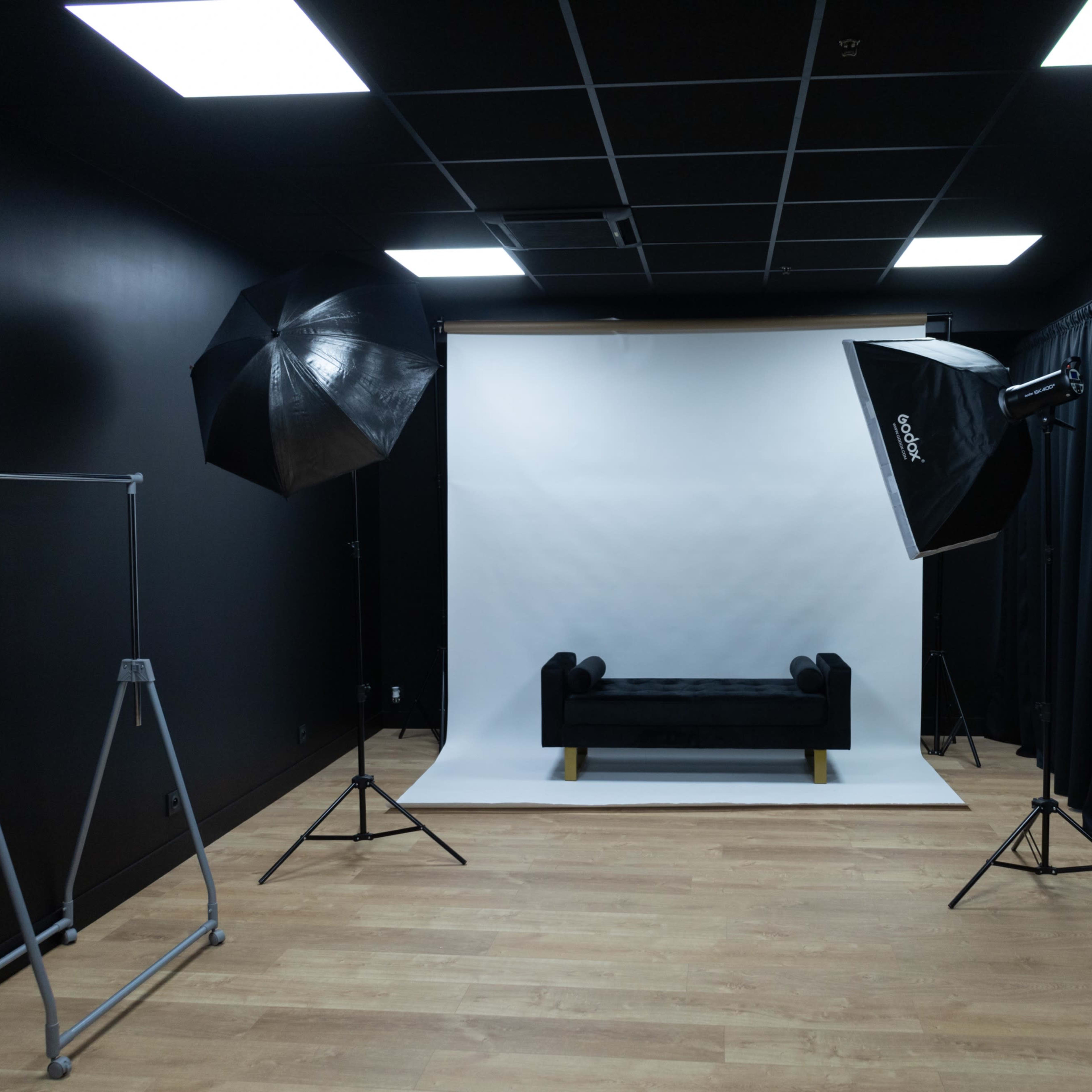 A photography studio with a black backdrop, a white backdrop, a black couch, lighting equipment, and a clothing rack.