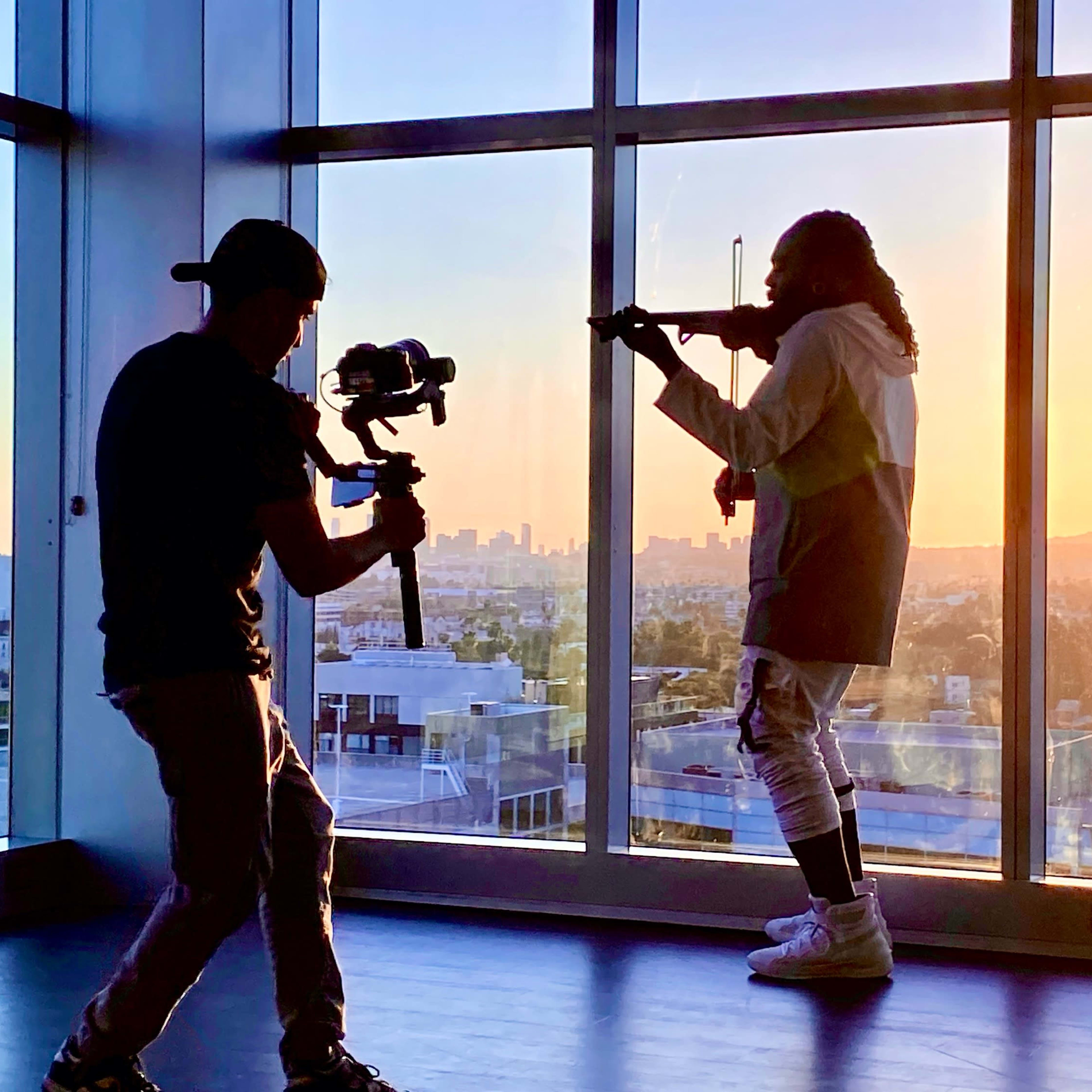 A silhouette of a musician playing the violin is captured against a sunset while a cameraman films the scene.