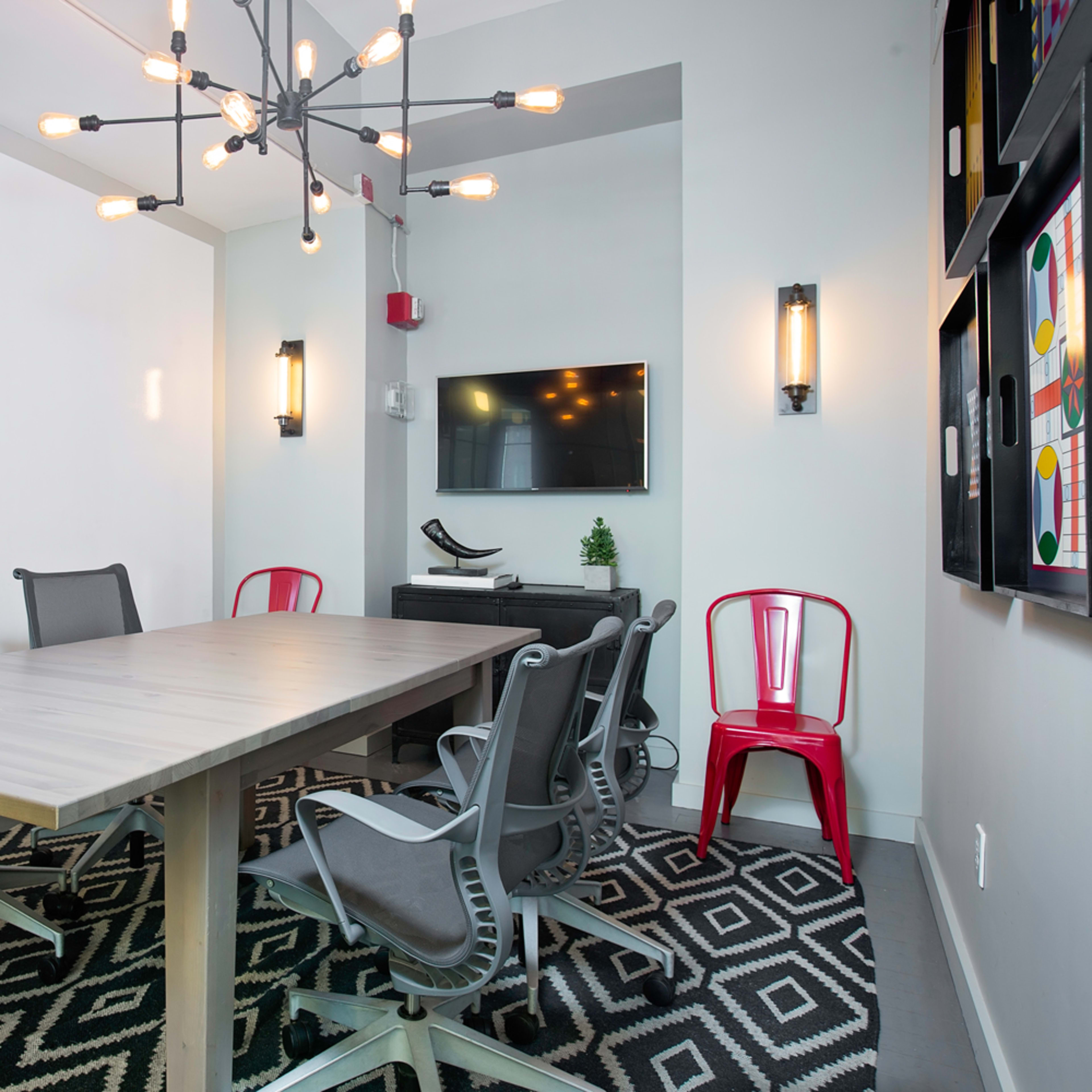 A modern conference room features a long wooden table surrounded by six gray chairs, a red chair, and a chandelier overhead, with a television mounted on the wall and glass-paneled doors.