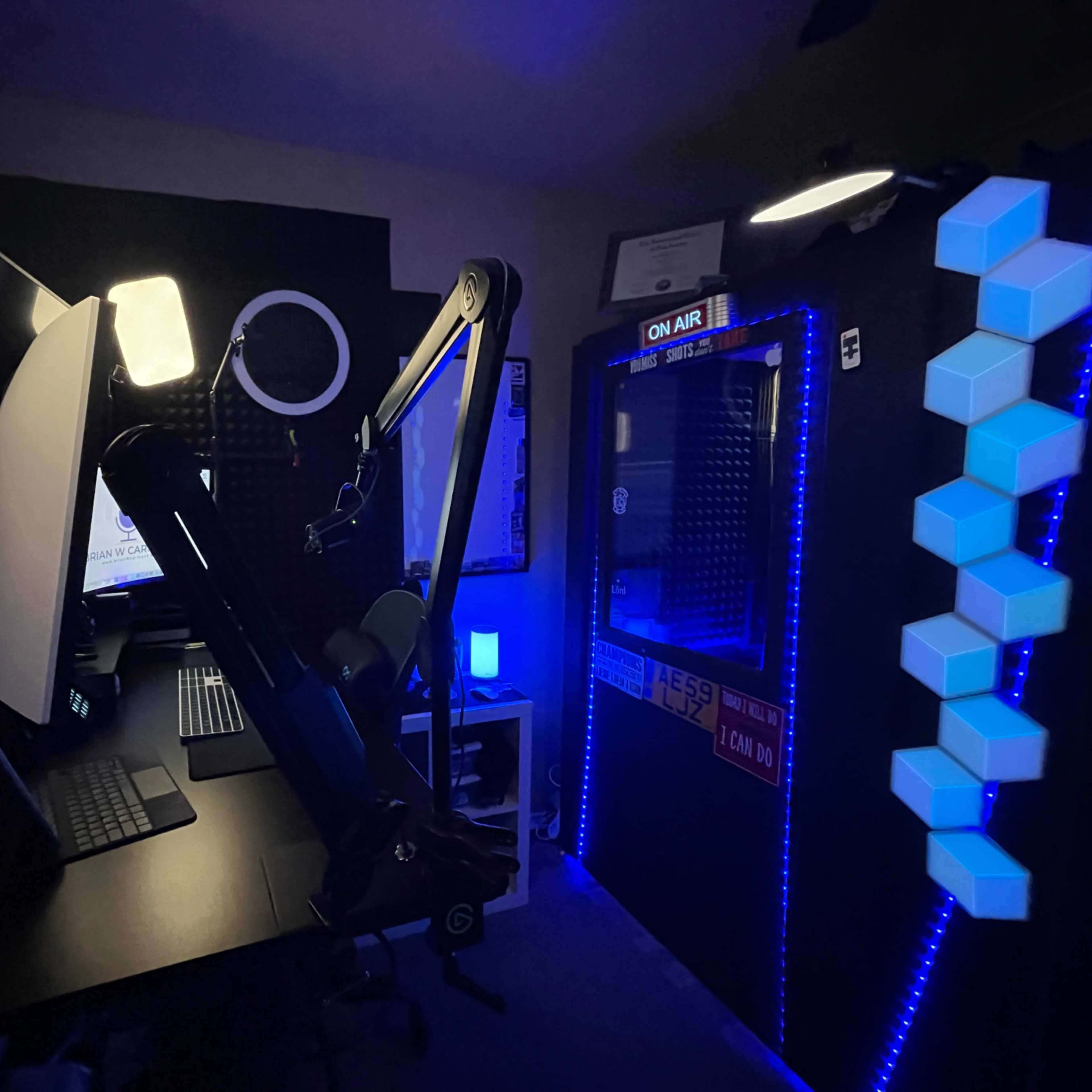 A modern workspace featuring a computer setup with multiple screens, a microphone on an adjustable arm, and decorative LED lights along a wall.