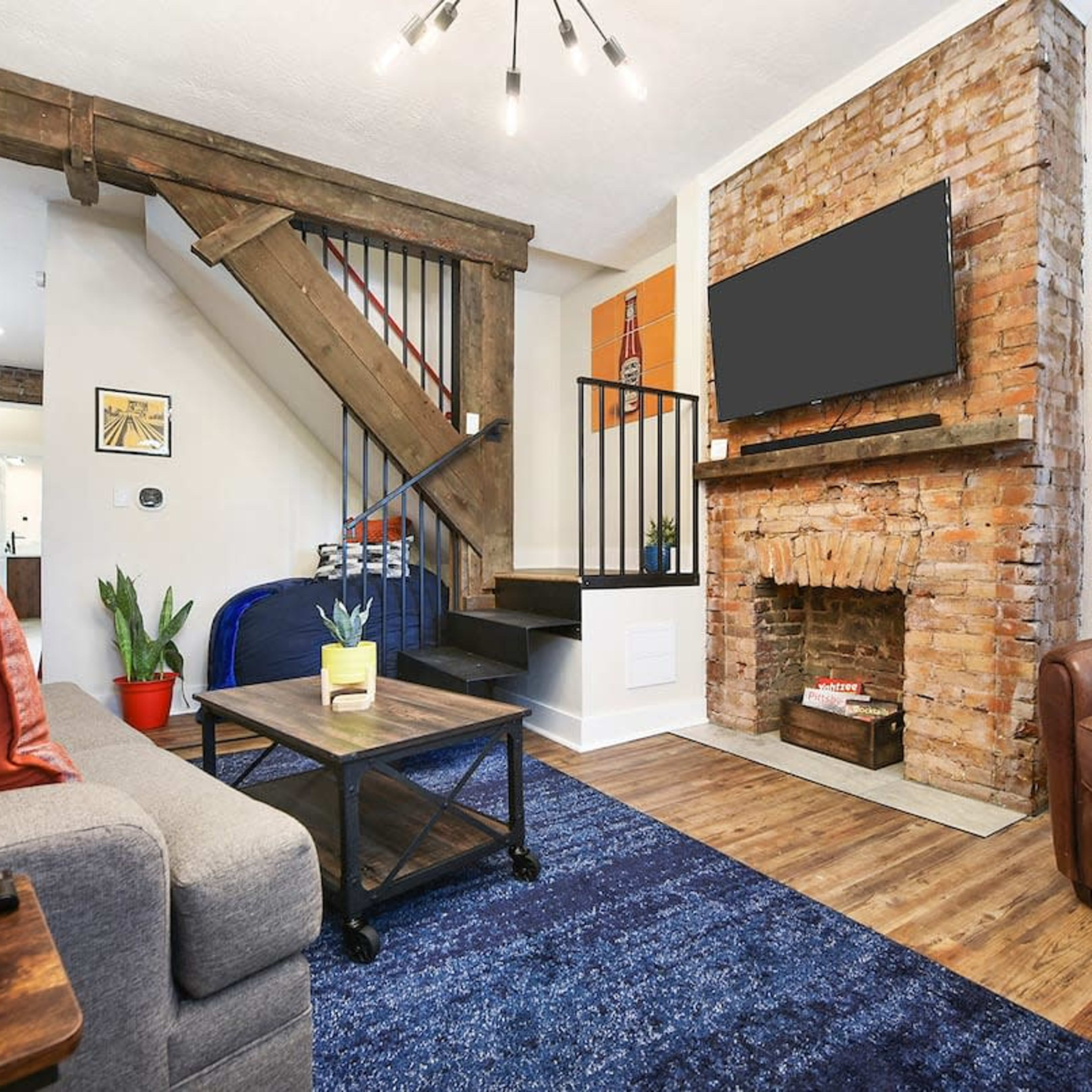 A cozy living room with a brick fireplace, a staircase to the upper level, and a television mounted on the wall, furnished with a sofa, armchair, and coffee table.