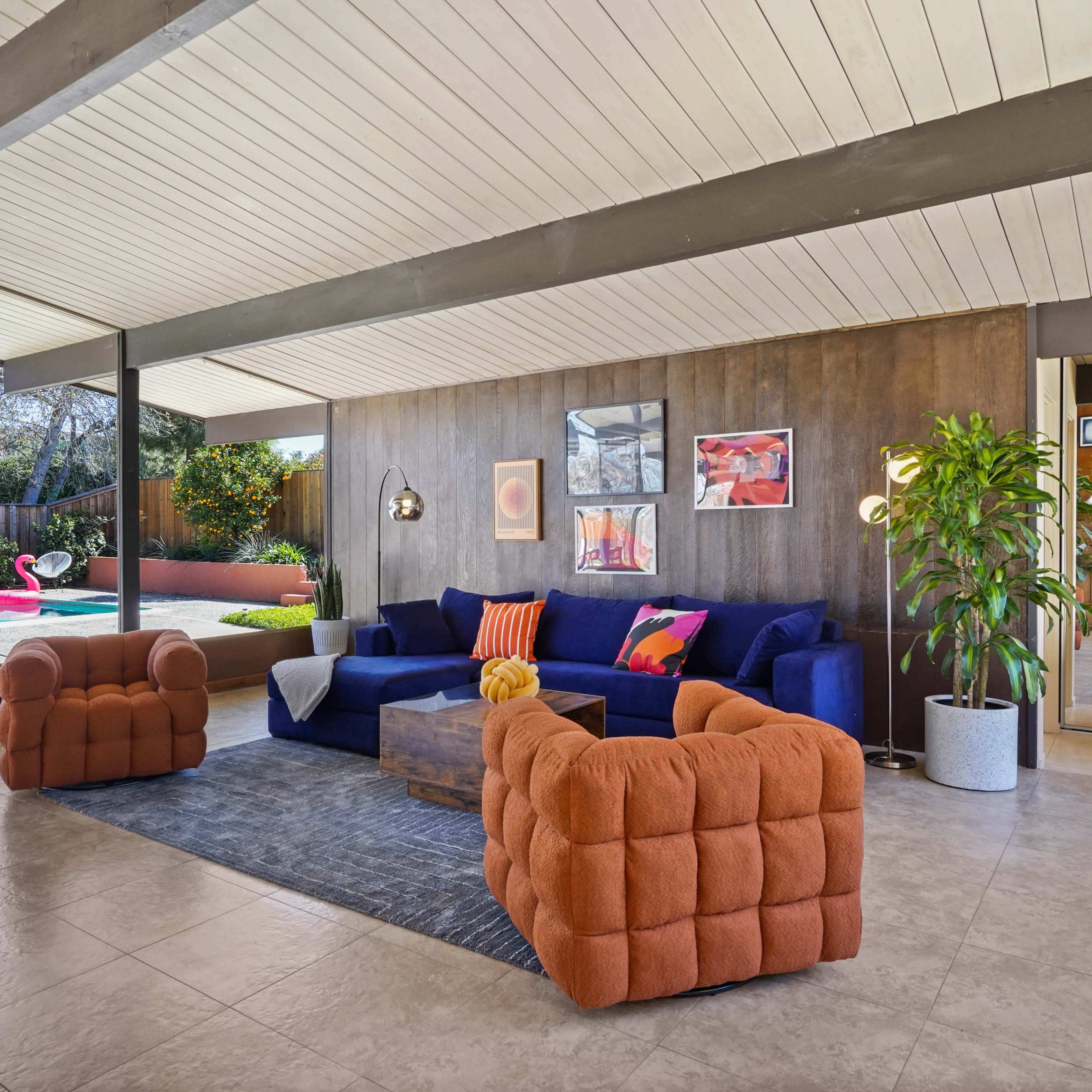 A modern living room features a blue sectional sofa, orange chairs, a coffee table, and large windows overlooking a pool area.