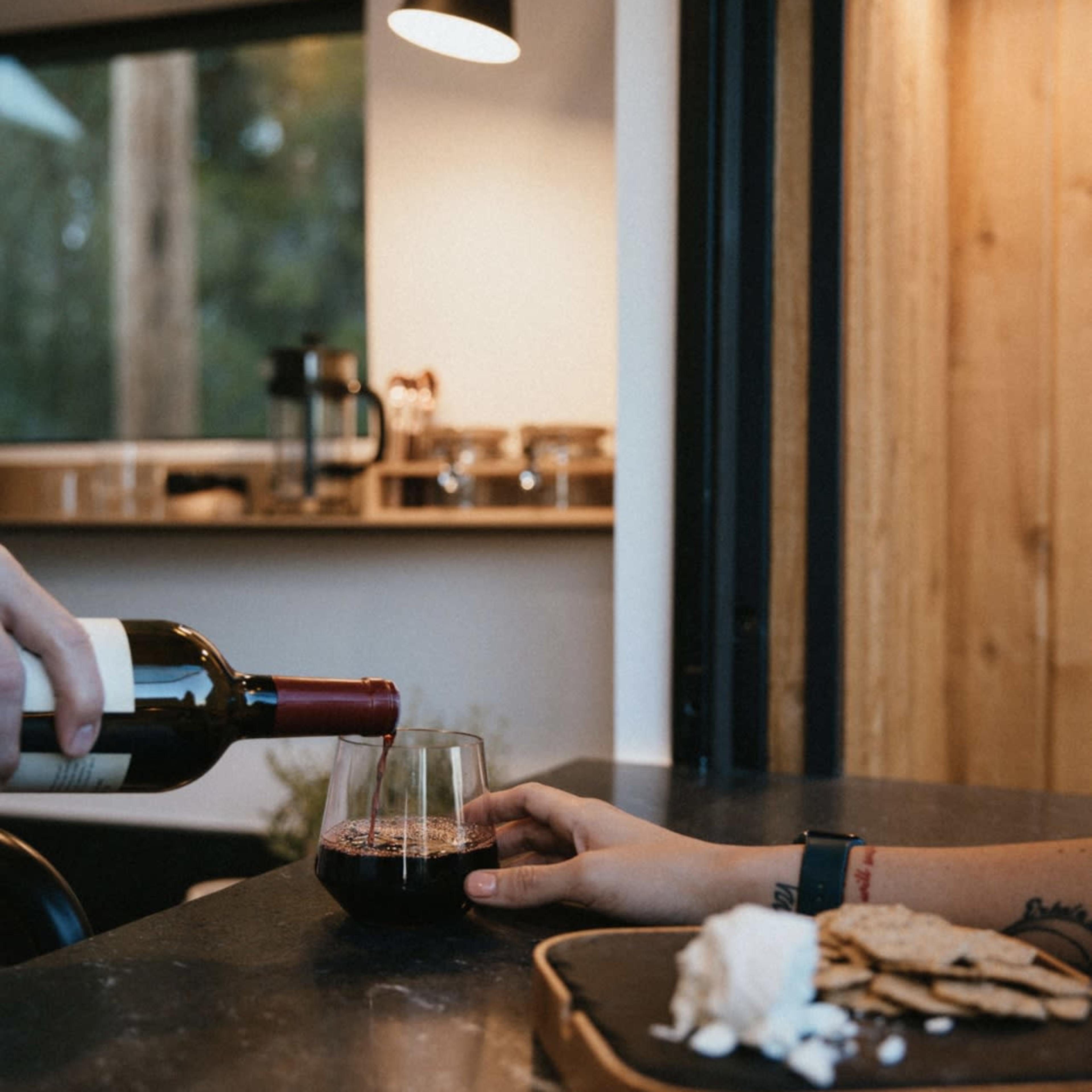 A hand pours red wine into a glass while another hand reaches for a plate of crackers and cheese on a table.