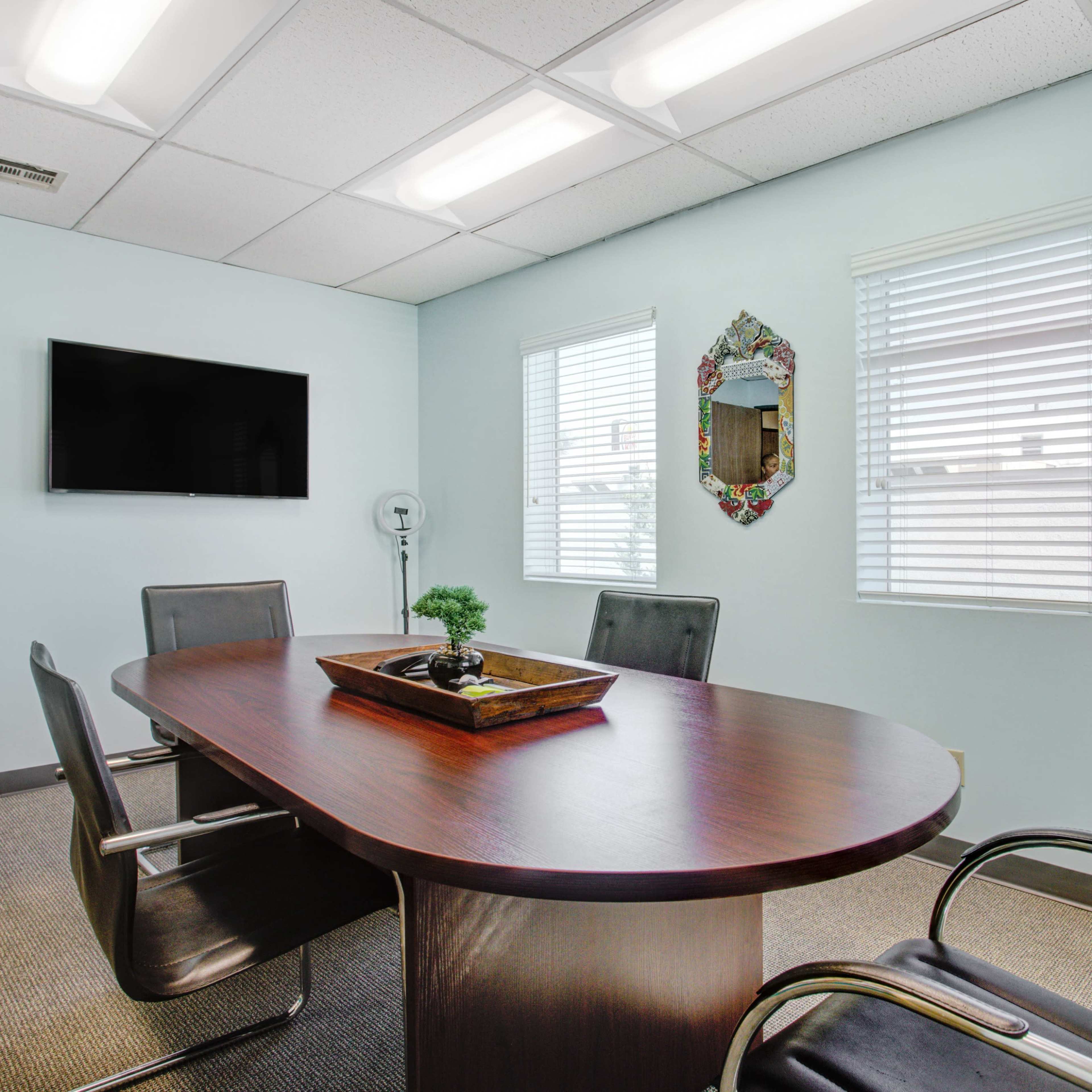 A conference room features a wooden table with a decorative tray, four black chairs, a wall-mounted TV, and two windows with blinds.