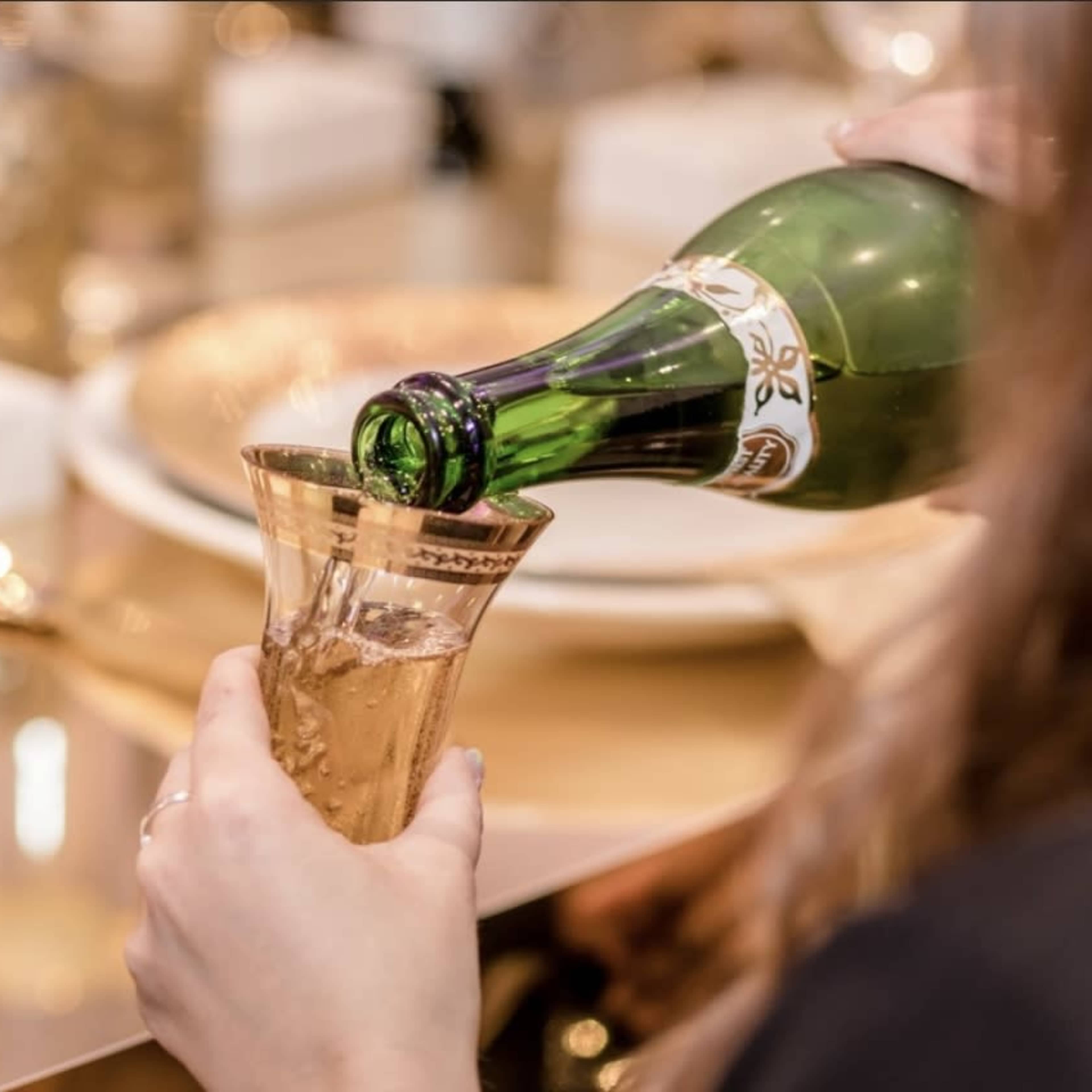 A hand pours sparkling wine into a decorative glass at a table set with fine china and silverware.