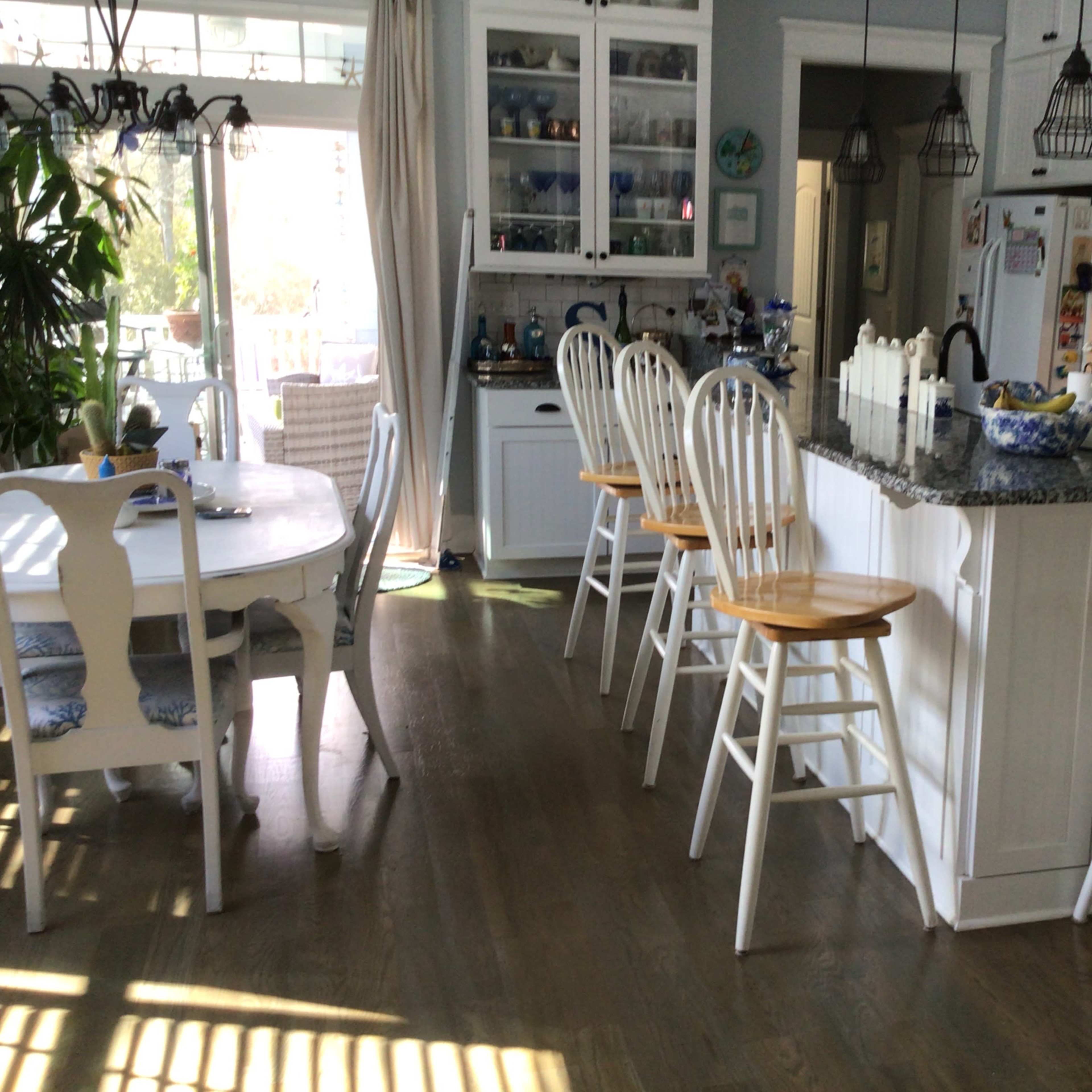 A bright kitchen with a granite countertop, white bar stools, and a dining table adjacent to it, featuring natural light streaming through large windows.