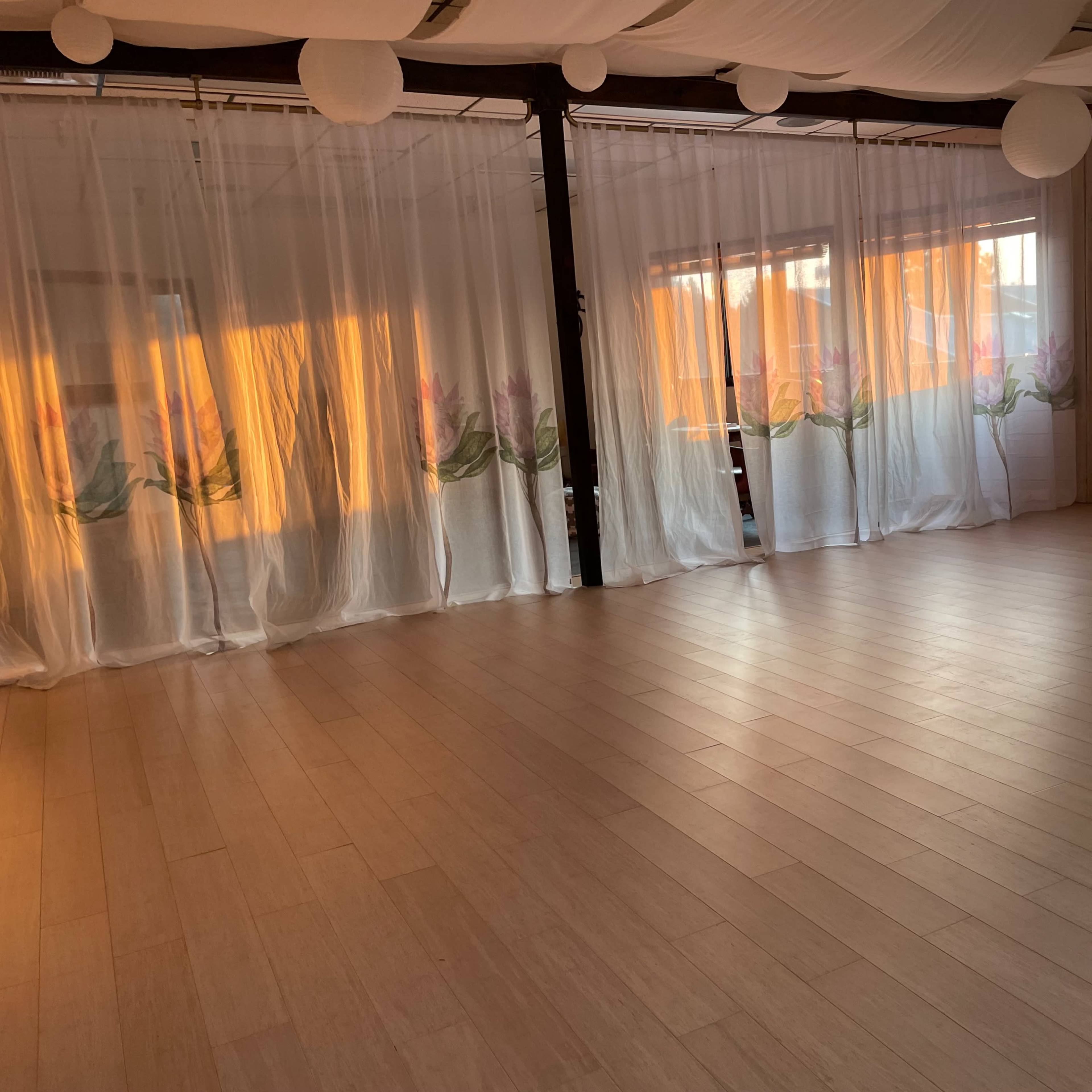 A spacious, light-filled room with wooden floors, sheer curtains featuring floral designs, and large windows allowing sunlight to stream in.