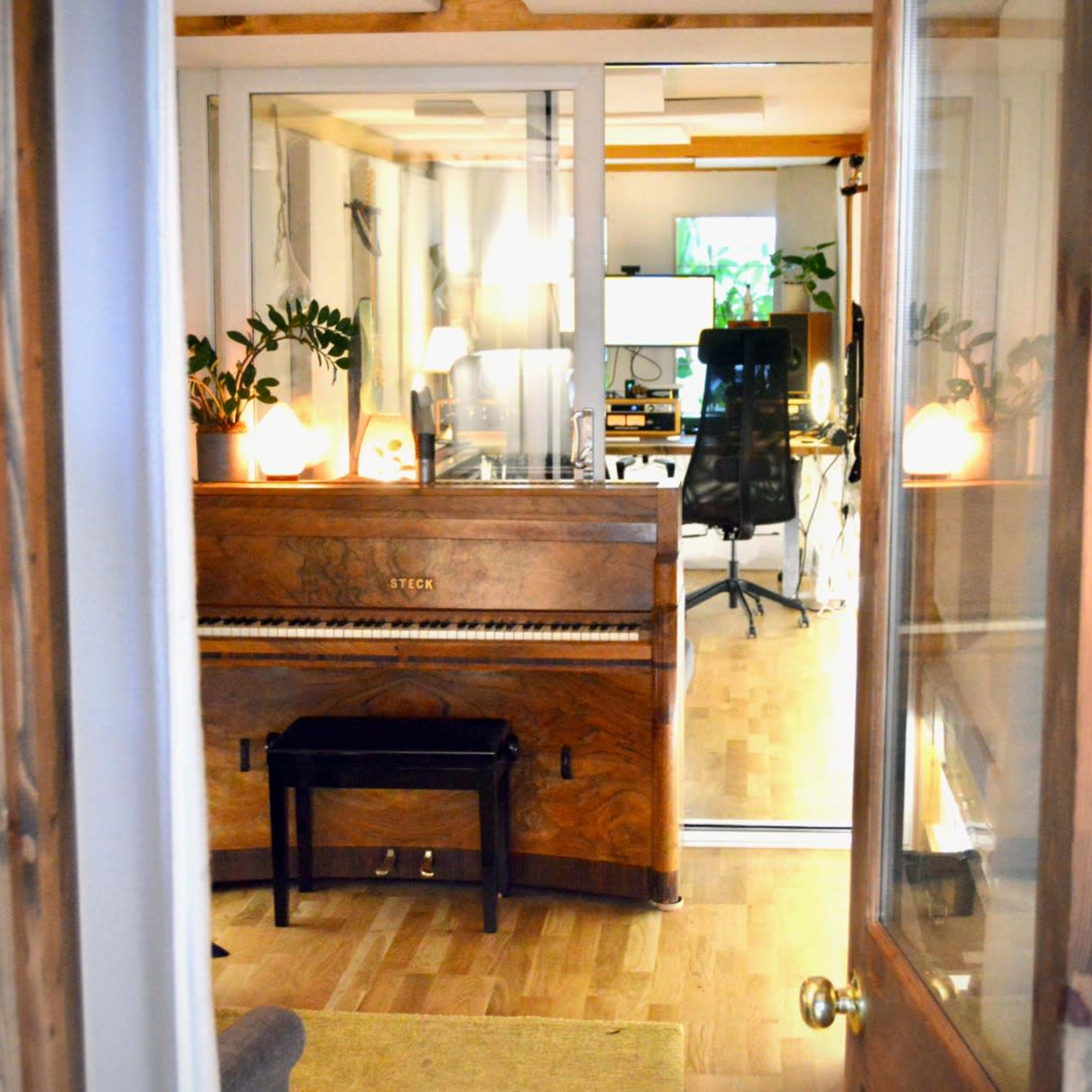 A wooden piano sits in a softly lit room, visible through a glass door that leads to a workspace with computers and plants.