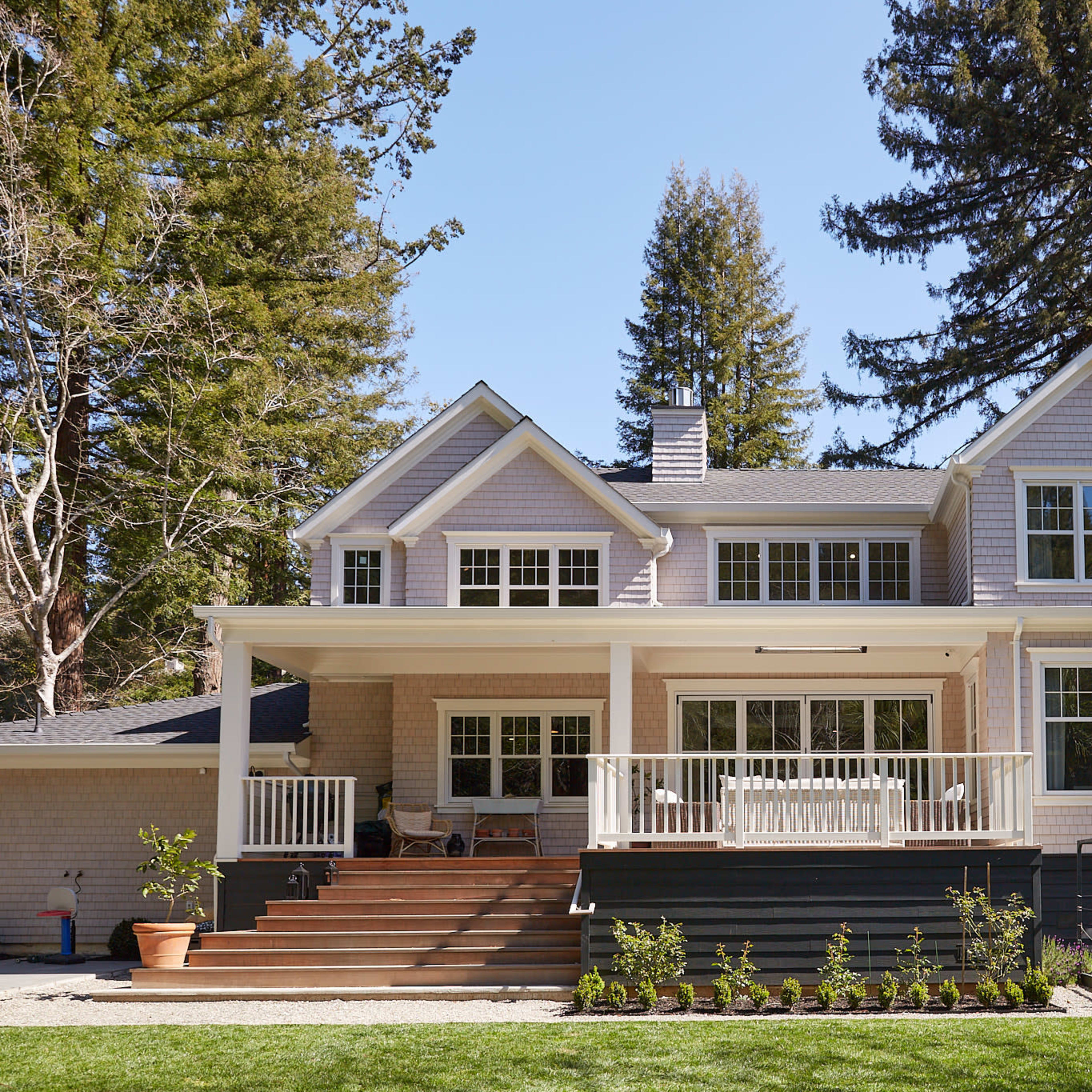 A large, light-colored house features multiple windows, a spacious deck with steps, and is surrounded by tall trees and a green lawn.