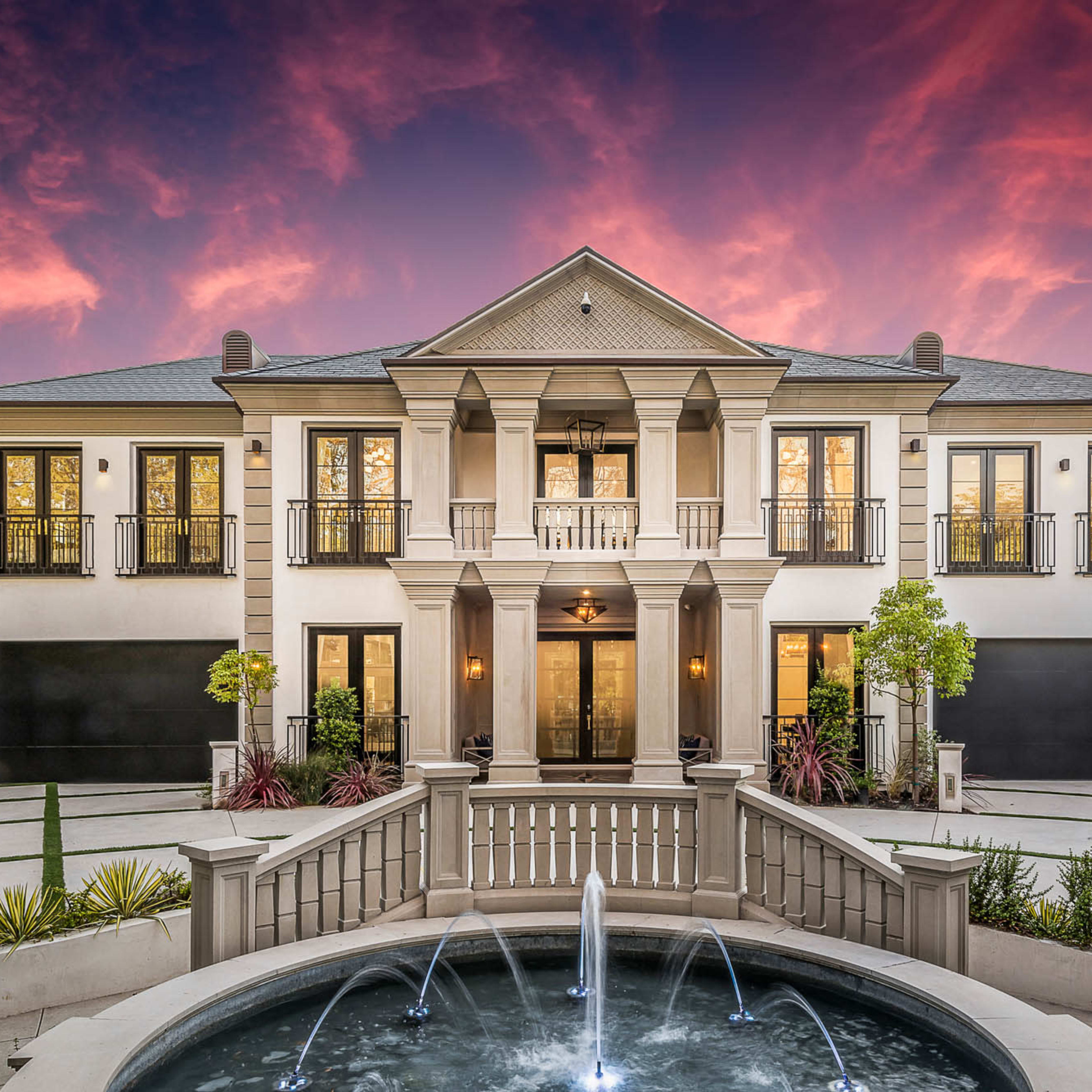 A large, two-story home features a grand entrance with multiple columns, a circular fountain in the foreground, and two garages, set against a vibrant sunset sky.