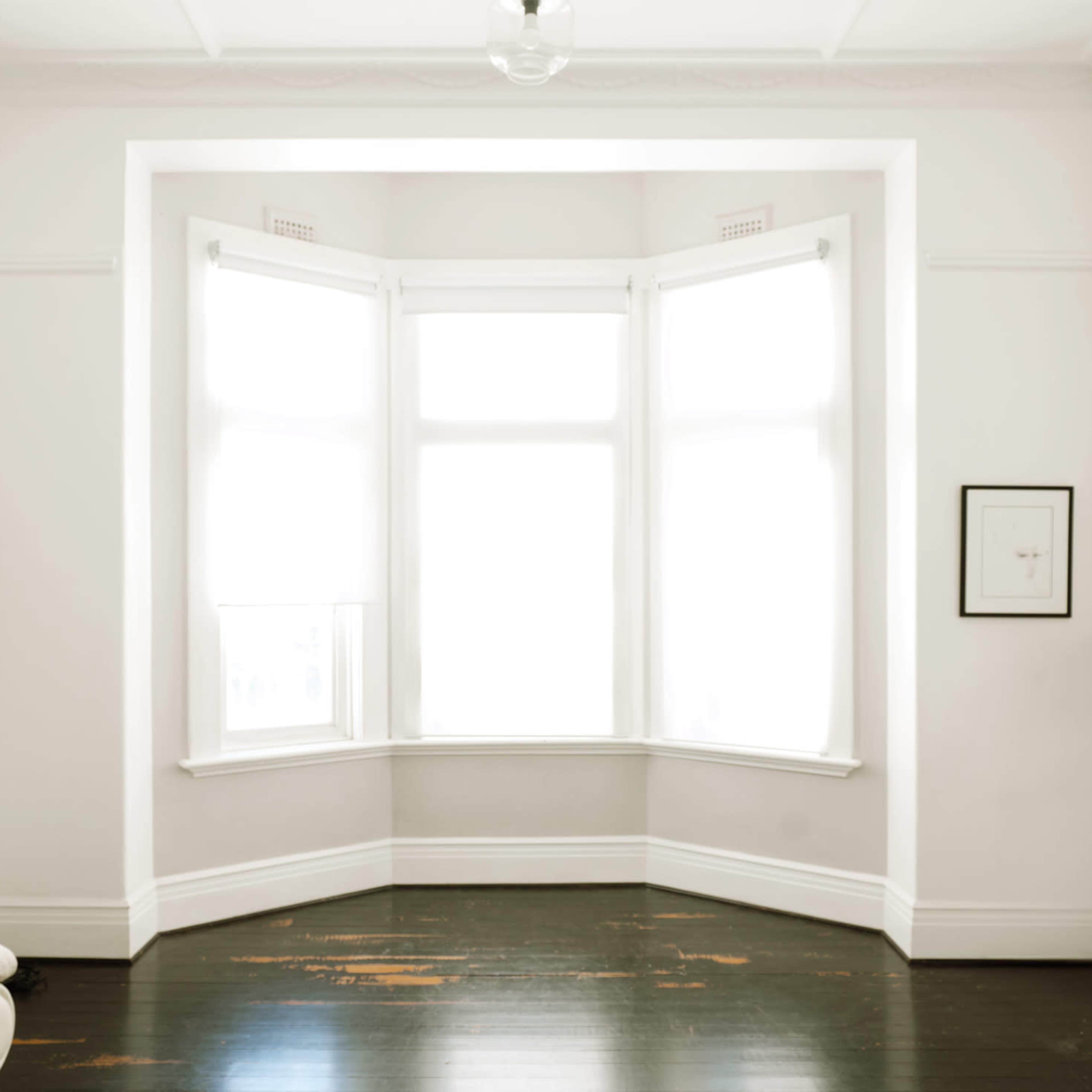 A bright, empty room features large windows with white curtains and dark wooden floors.
