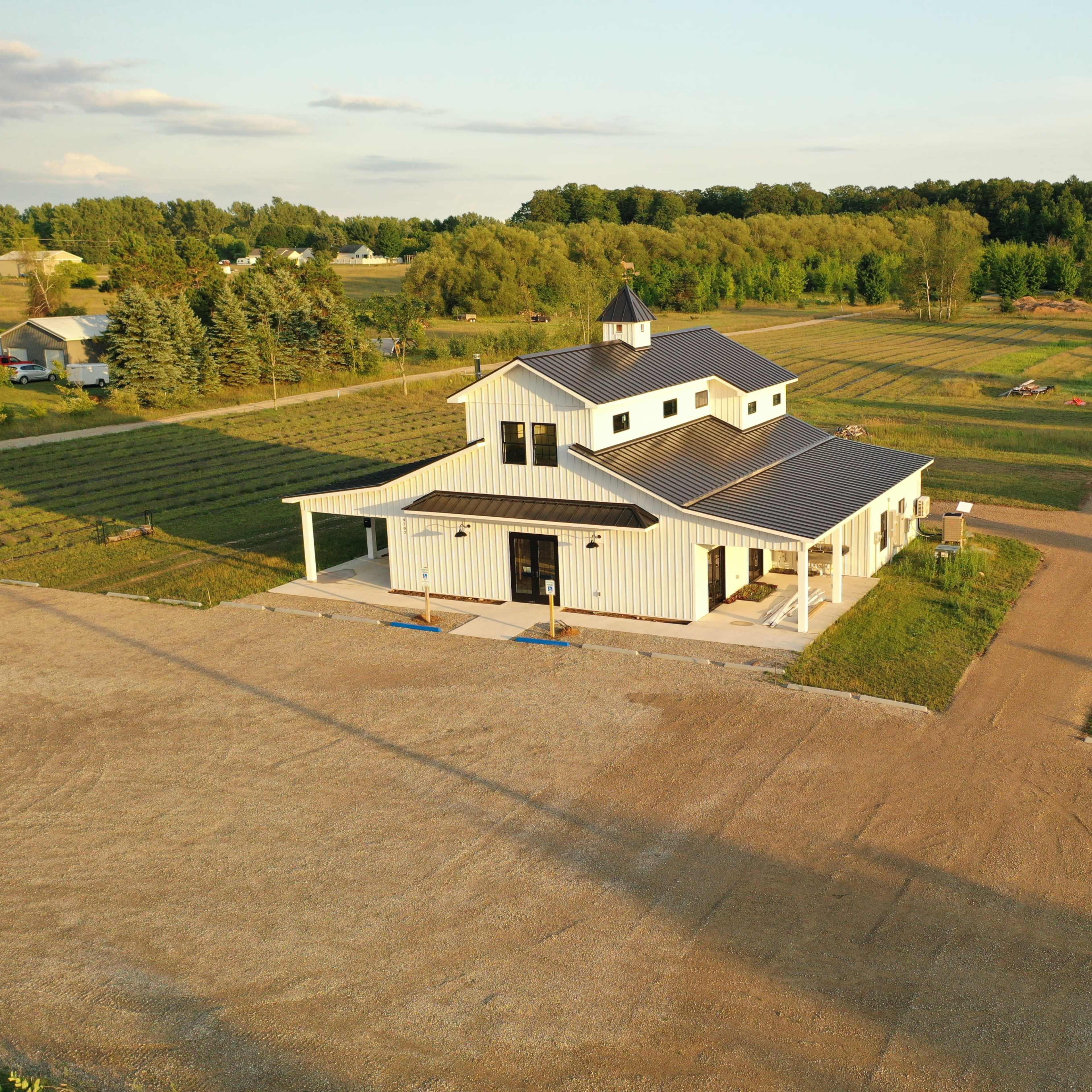 A modern farmhouse with a metal roof is situated on a gravel driveway, surrounded by fields and trees.
