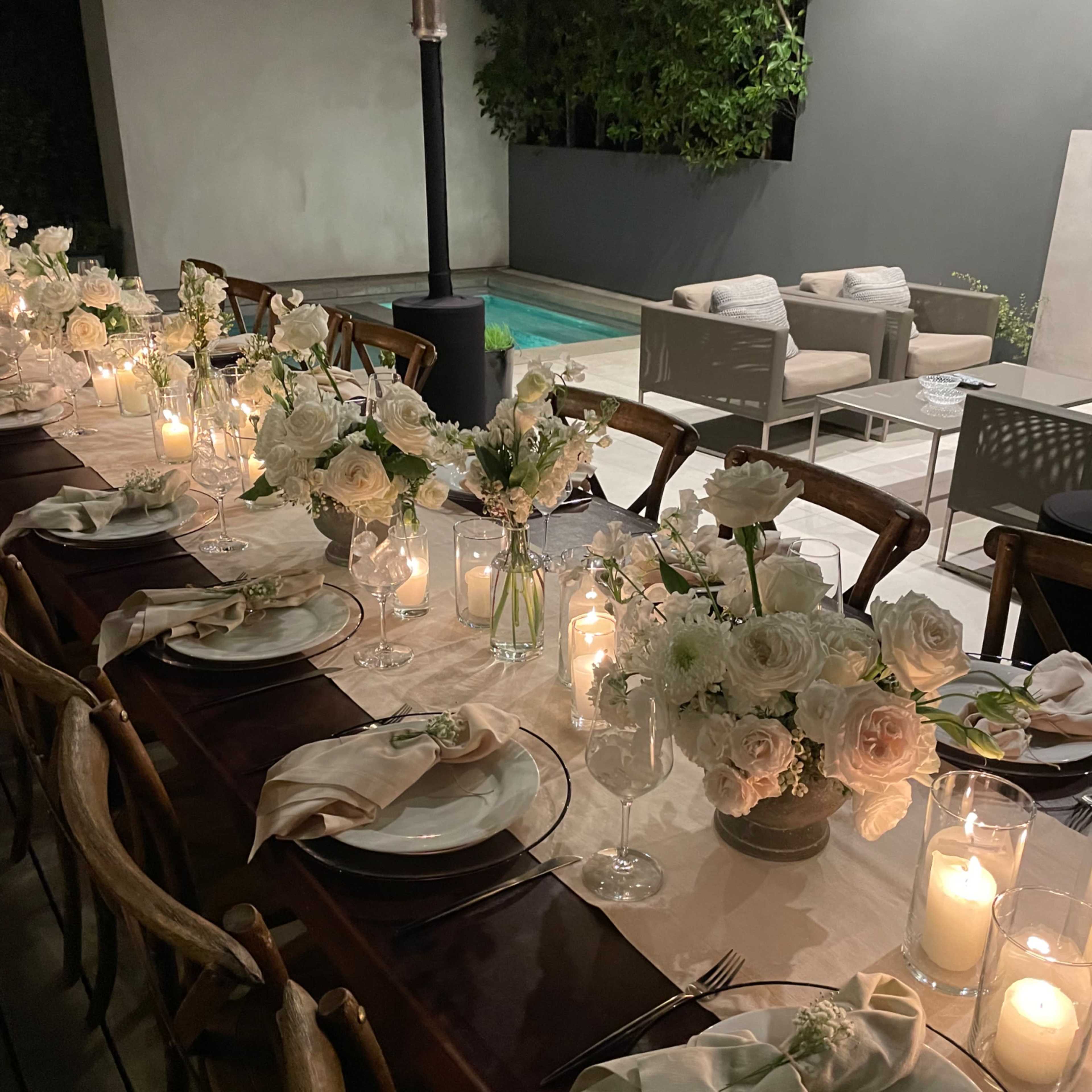A long dining table is set outdoors with elegant flower arrangements, candles, and plates, while a pool and seating area are visible in the background.