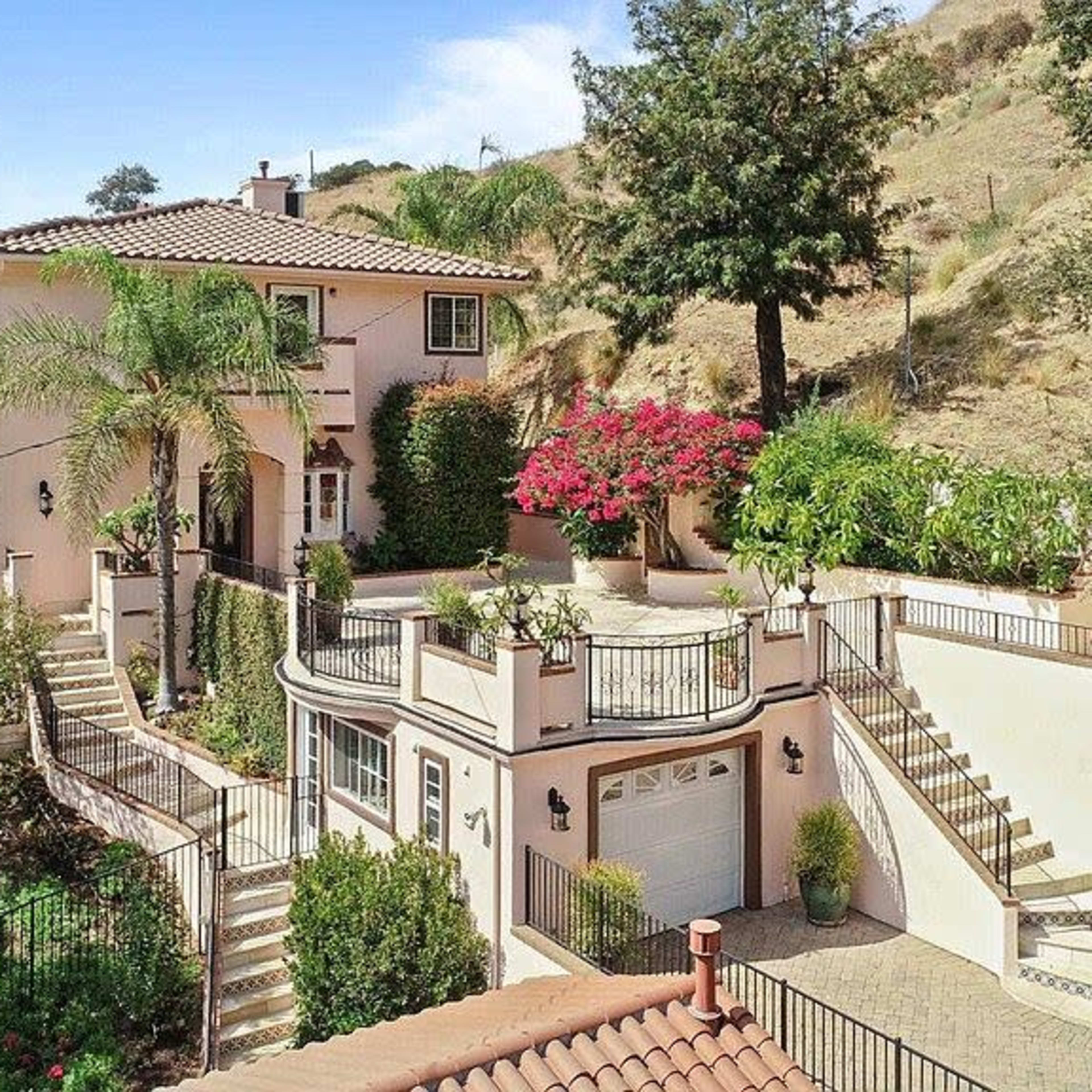A two-story pink house with a tile roof, surrounded by landscaped gardens and palm trees, situated on a hillside.