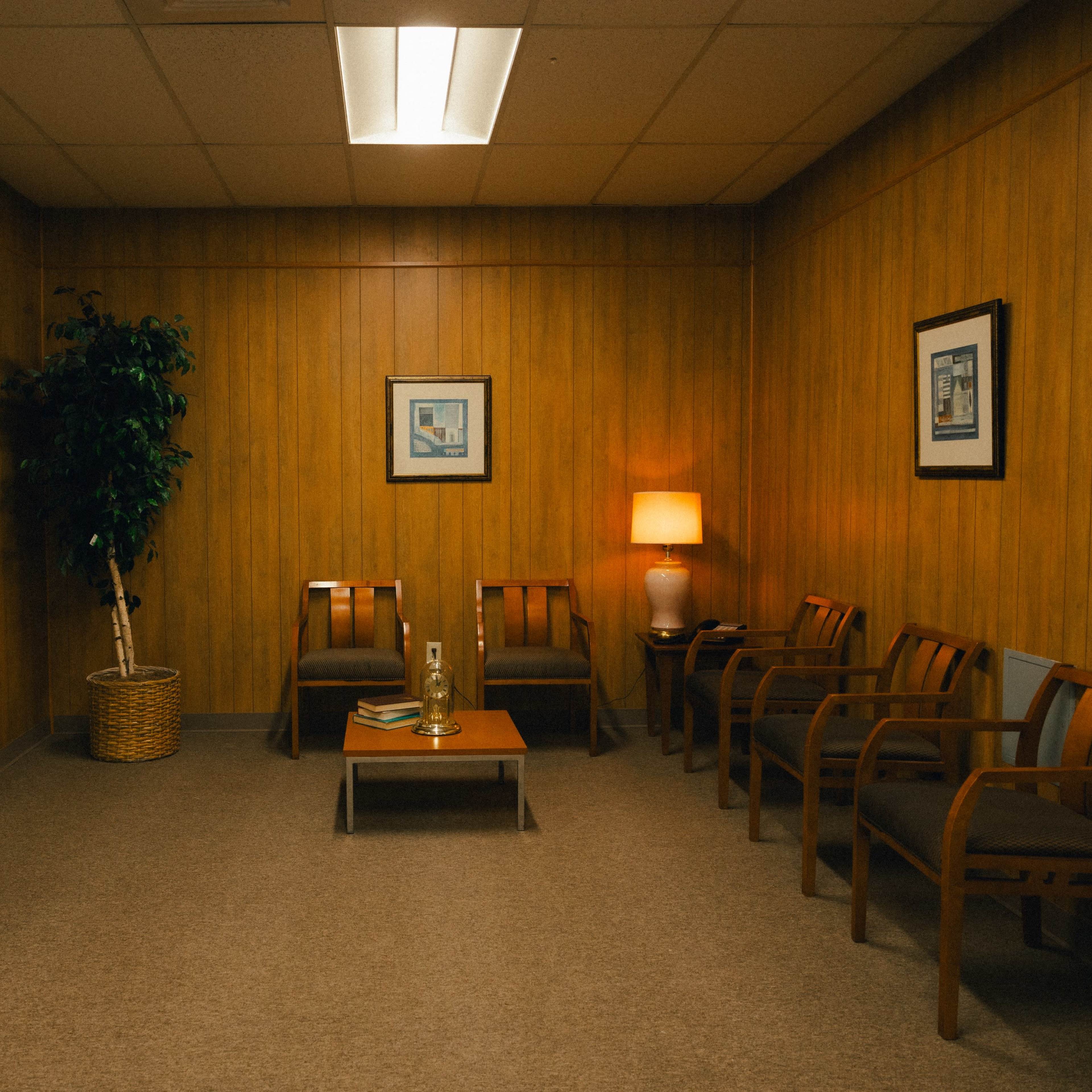 A waiting room features wooden paneling, two chairs, a small table with decorative items, a water cooler, and a lamp.