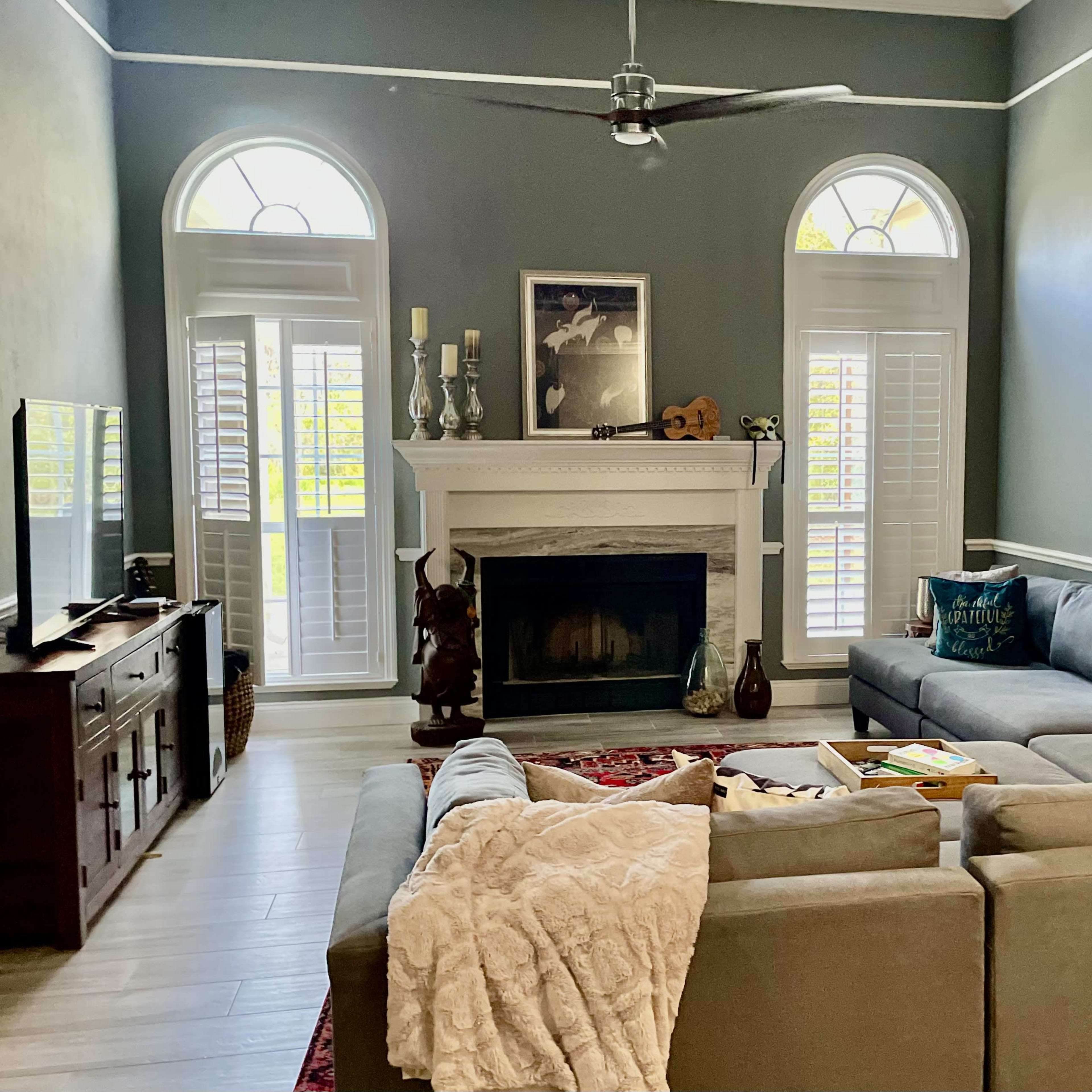 The living room features a large sectional sofa, a fireplace with decorative items, and tall windows with shutters.