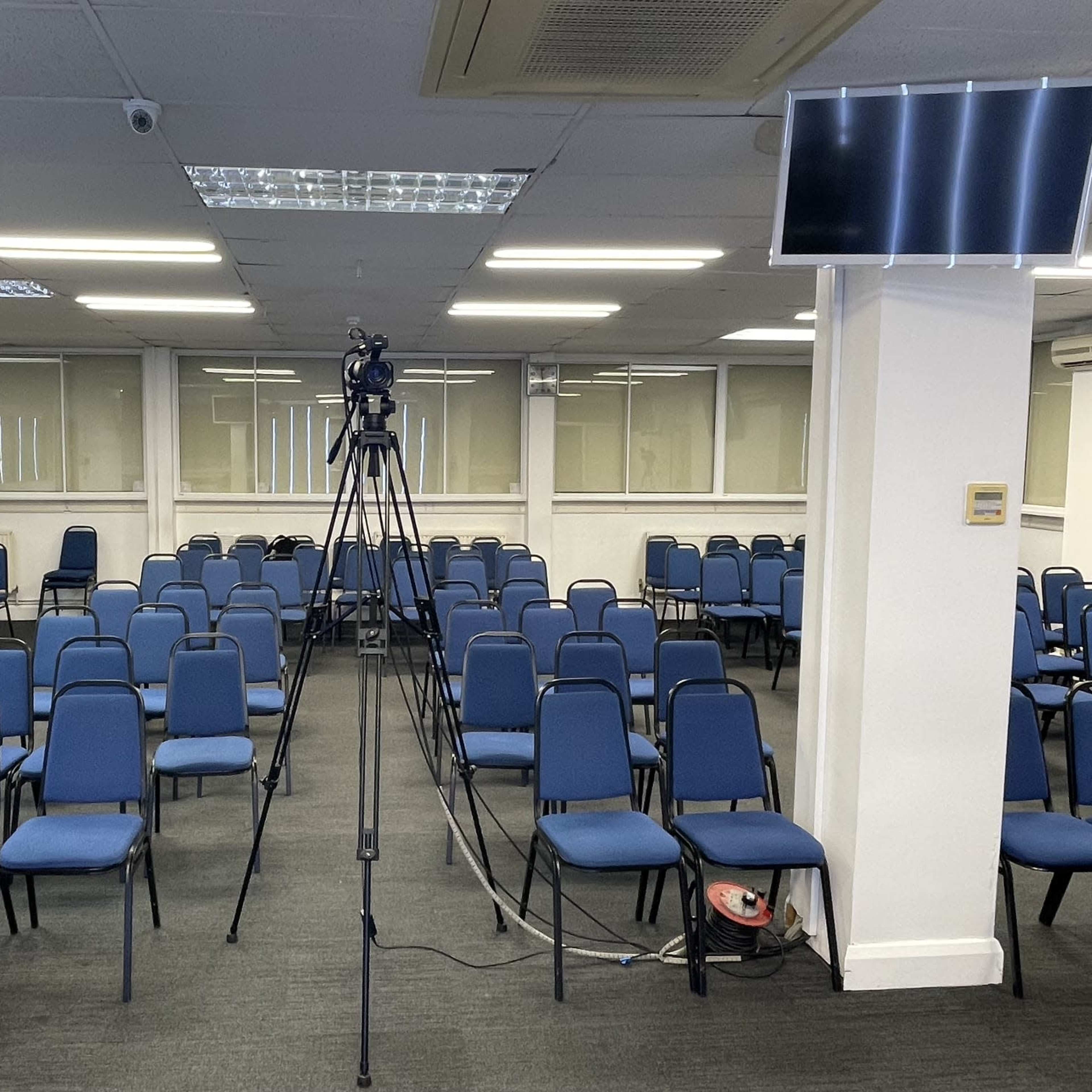 A large room filled with blue chairs arranged in rows, centered around a camera mounted on a tripod.