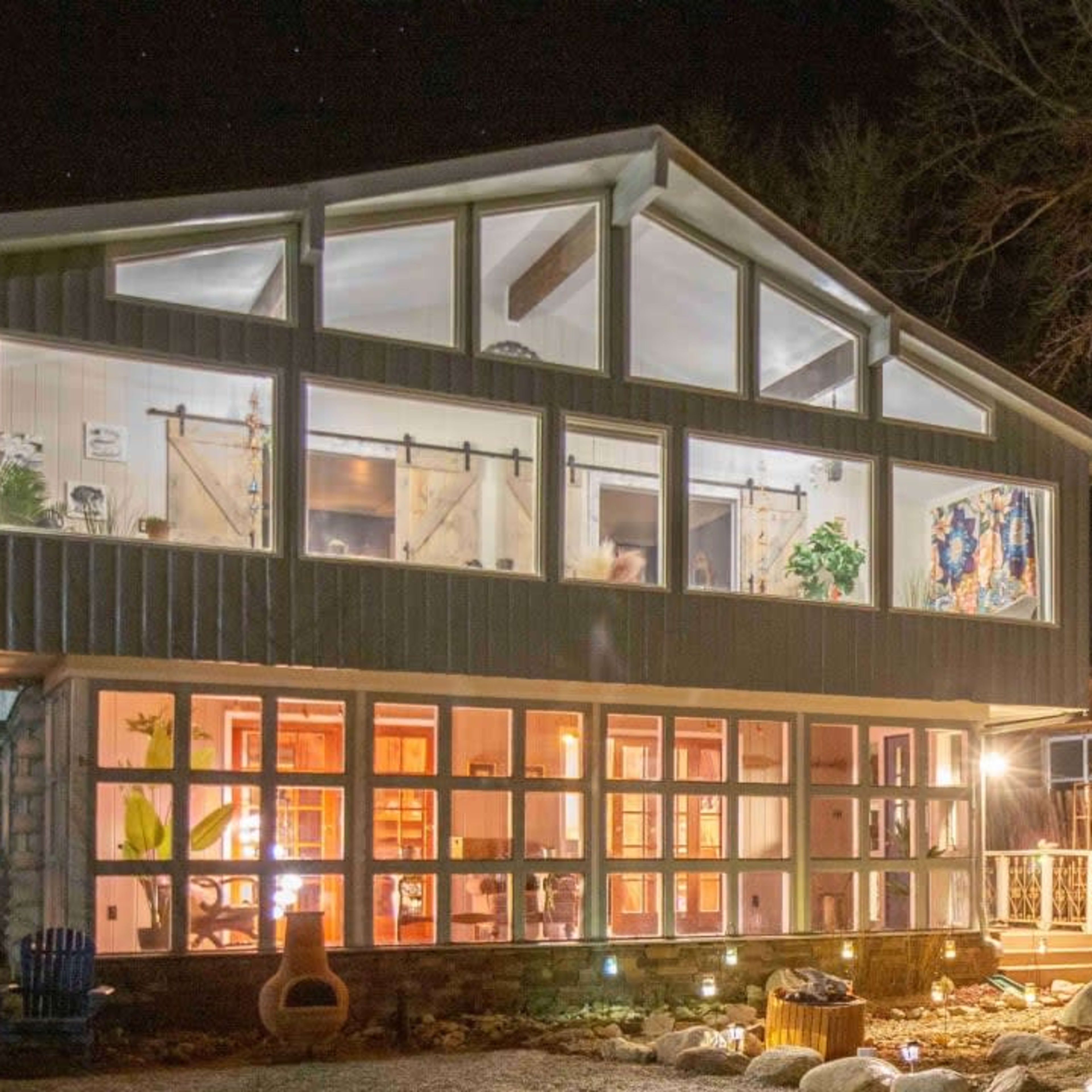 A modern two-story house with large windows, featuring a stone pathway and outdoor seating, is illuminated at night.