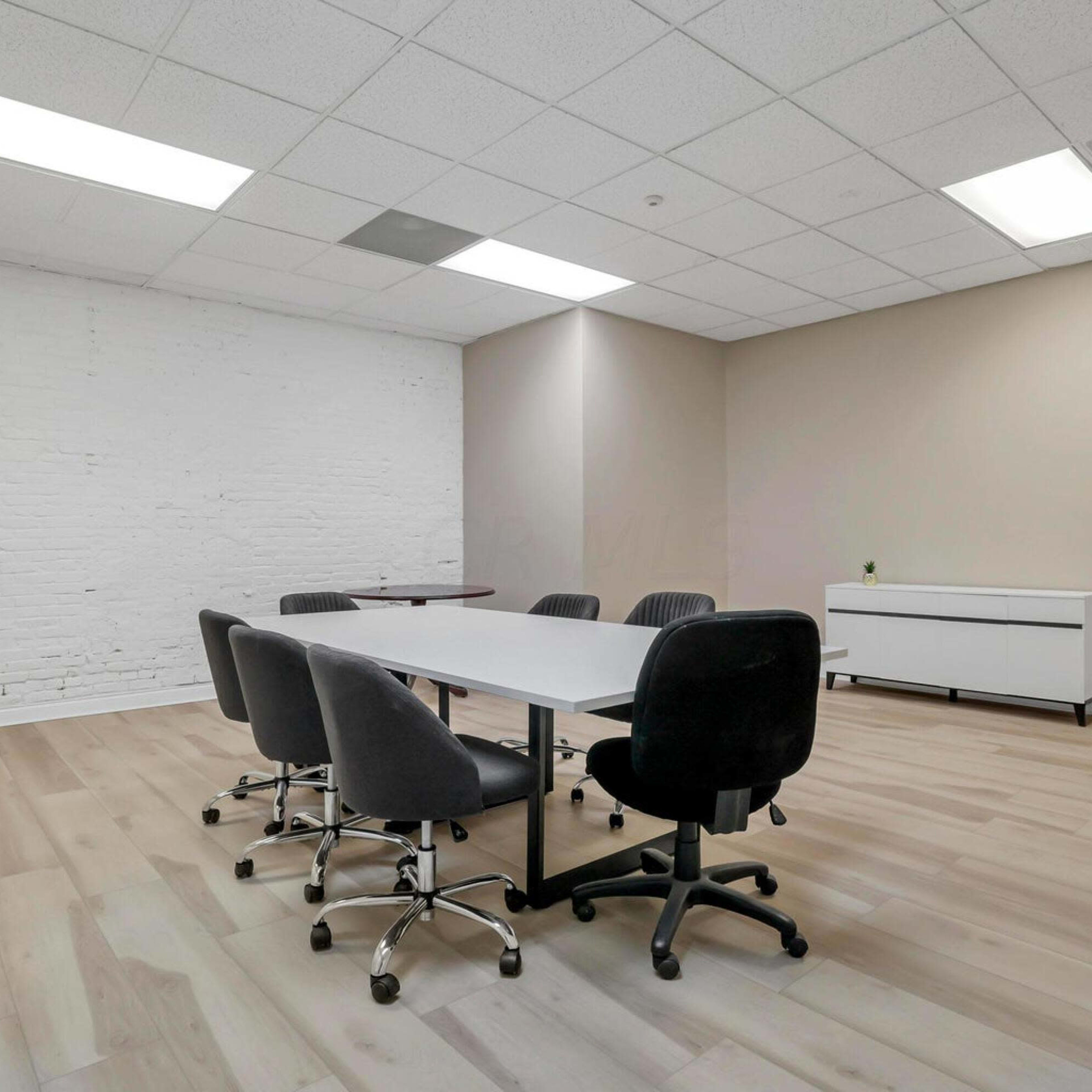A conference room features a rectangular table surrounded by six chairs, with a white brick wall and a cabinet in the background.