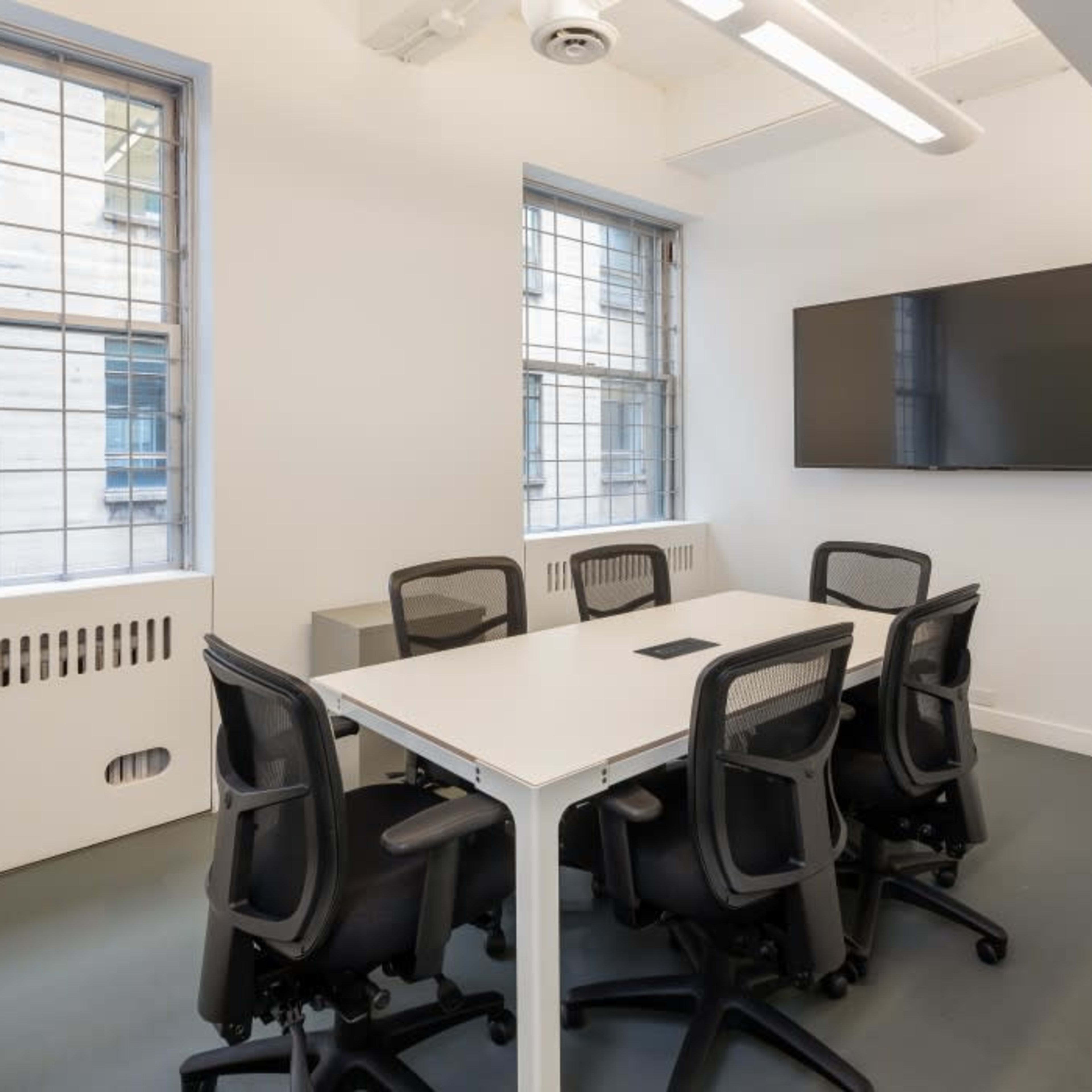 A modern conference room features a white table surrounded by black chairs, with a wall-mounted television and large windows providing natural light.
