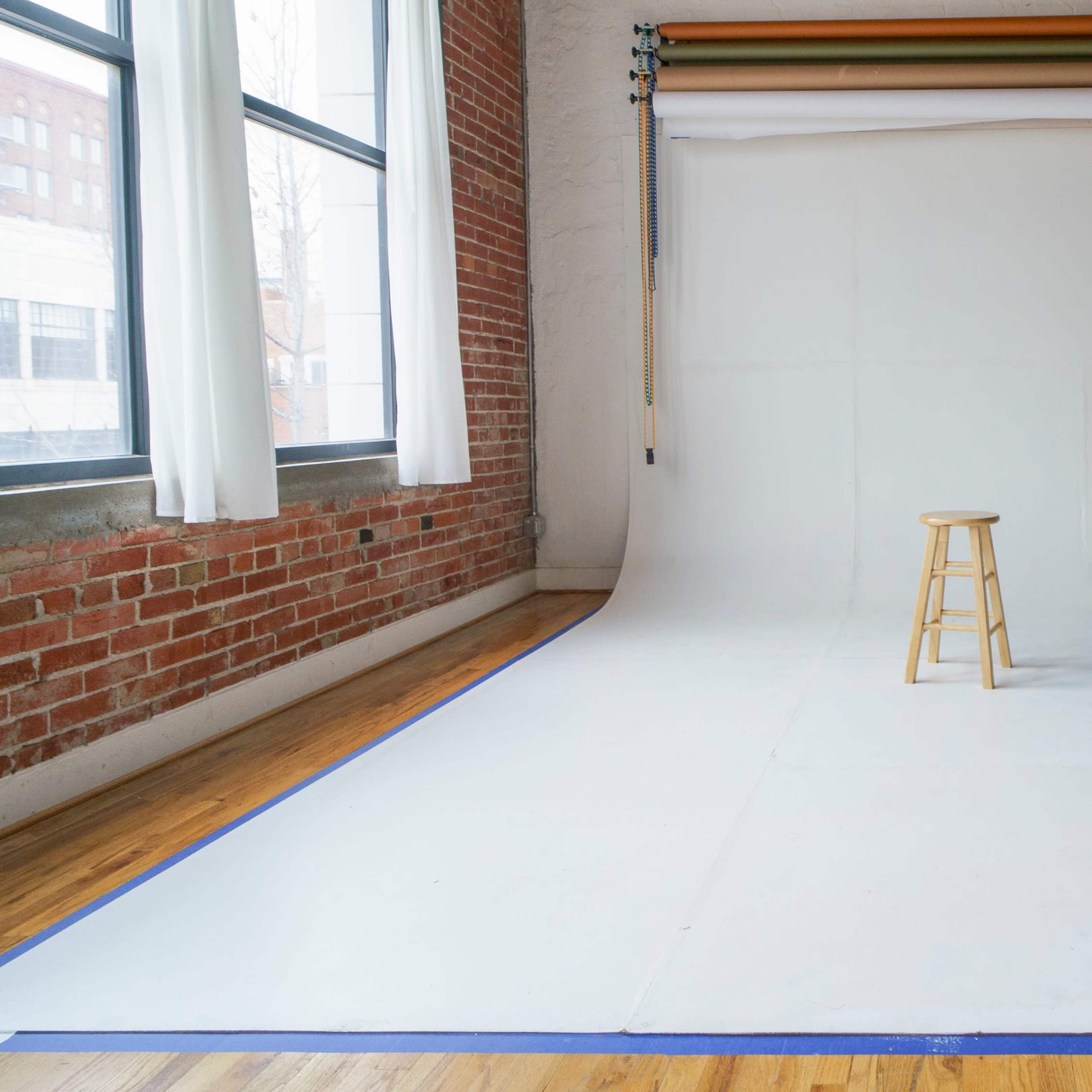 A photography studio with a white backdrop, a wooden stool in the center, and a bed in the background.