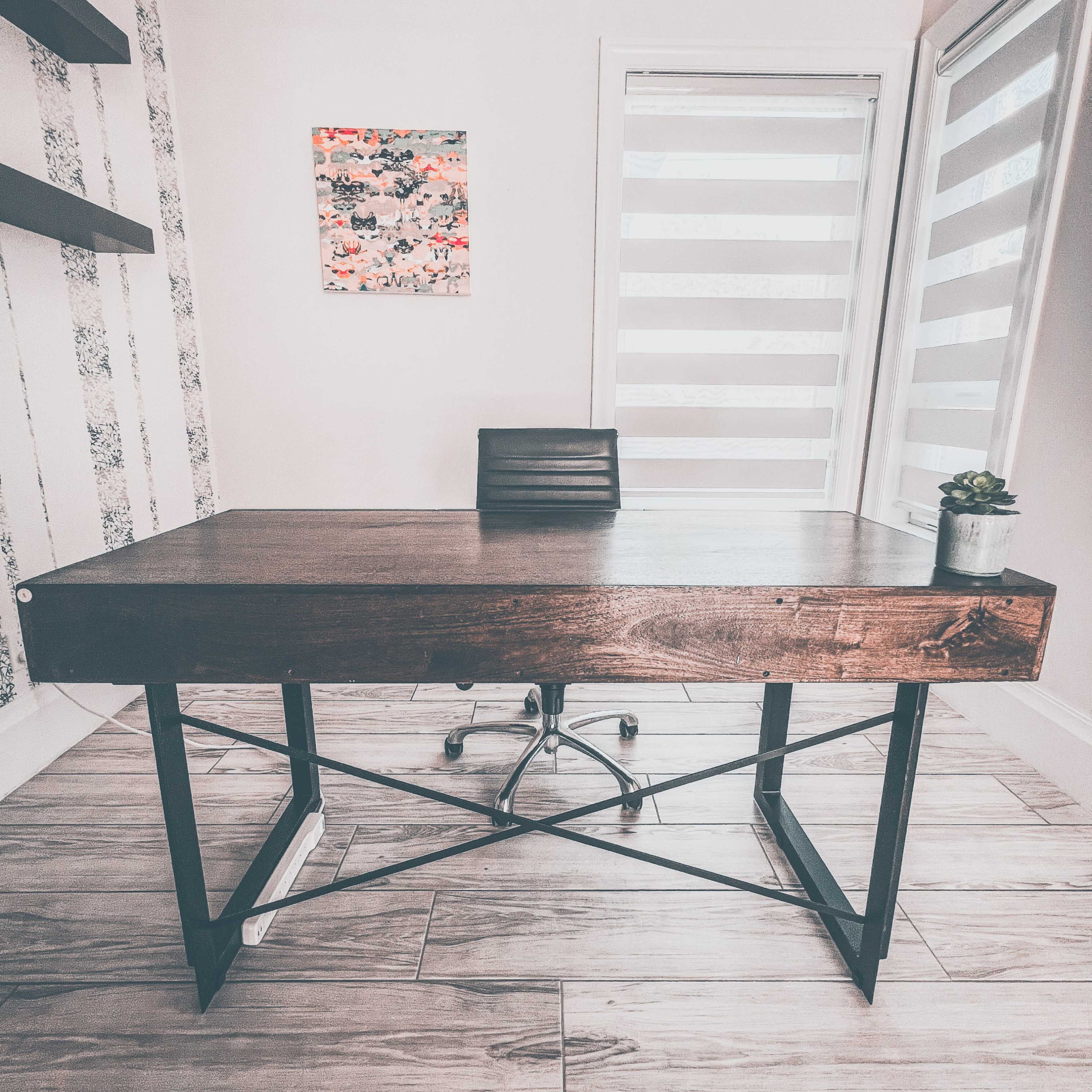 A modern office space features a wooden desk with a metal frame, a black ergonomic chair, a potted plant, and a piece of artwork on the wall.