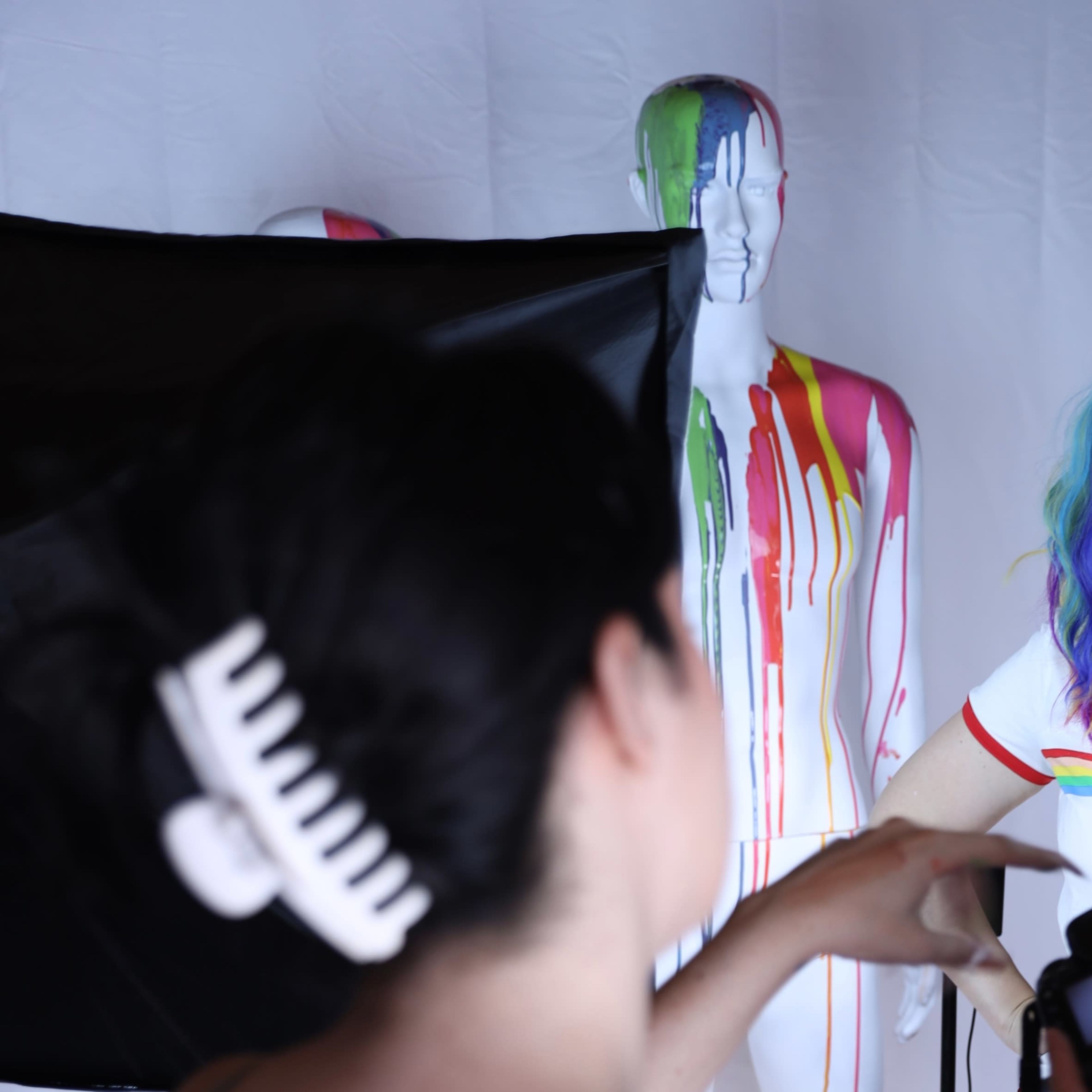 A woman with vibrant rainbow hair poses in front of a mannequin covered in colorful paint, while two people work on a photoshoot setup.