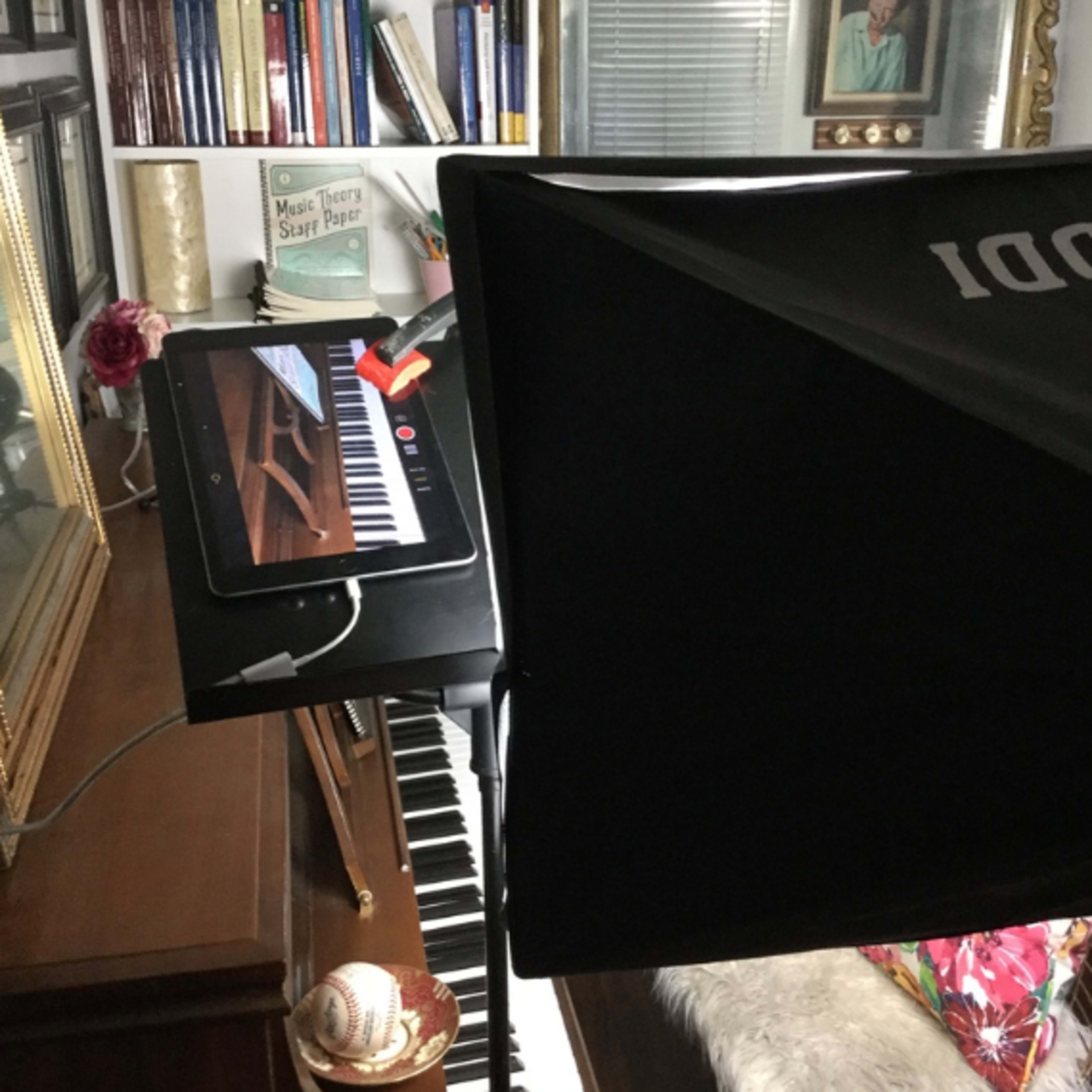 A piano with an iPad on a stand, illuminated by a softbox light in a room decorated with books, a framed photograph, and a sailboat model.