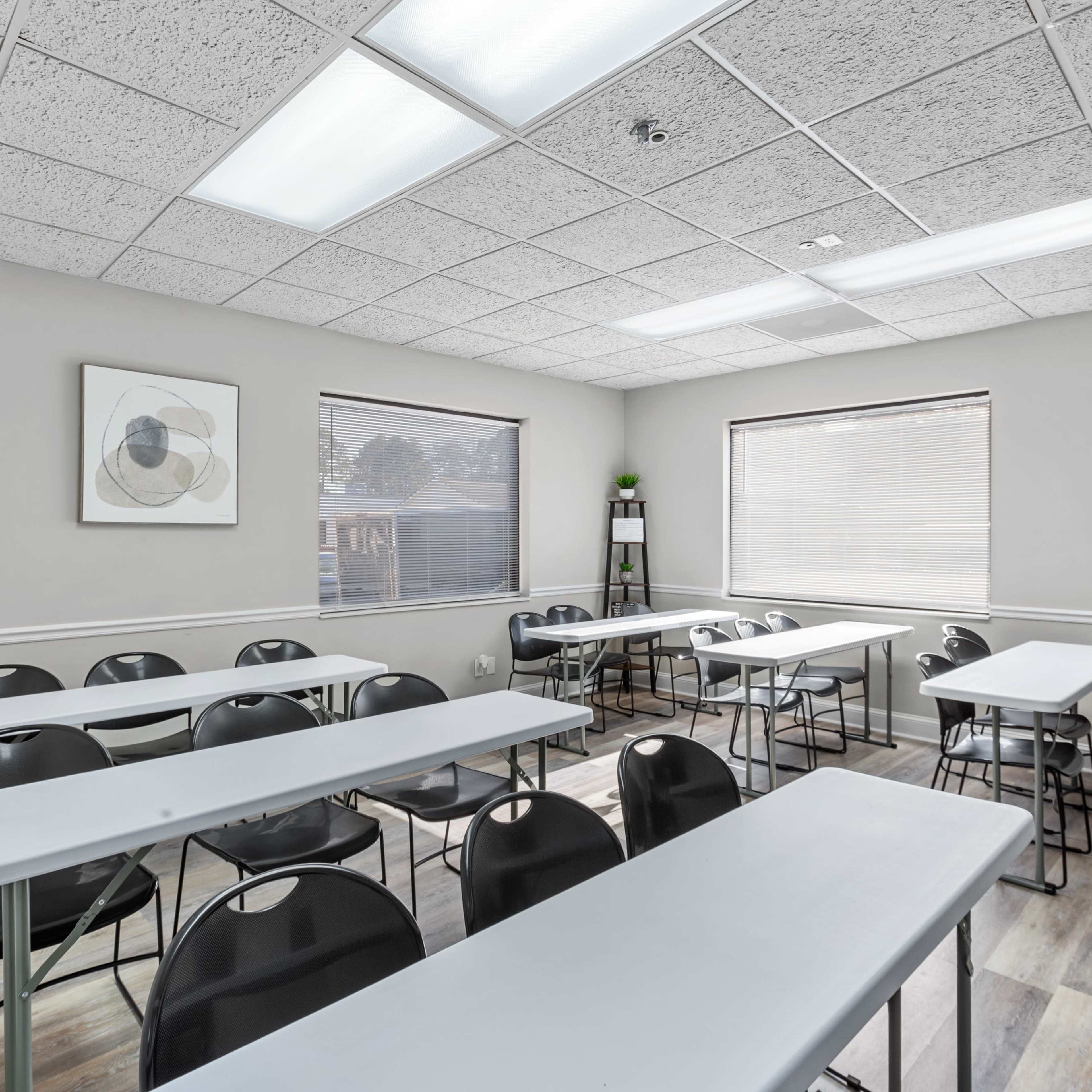 A classroom is set up with rows of tables and chairs, featuring large windows and a neutral-colored wall.