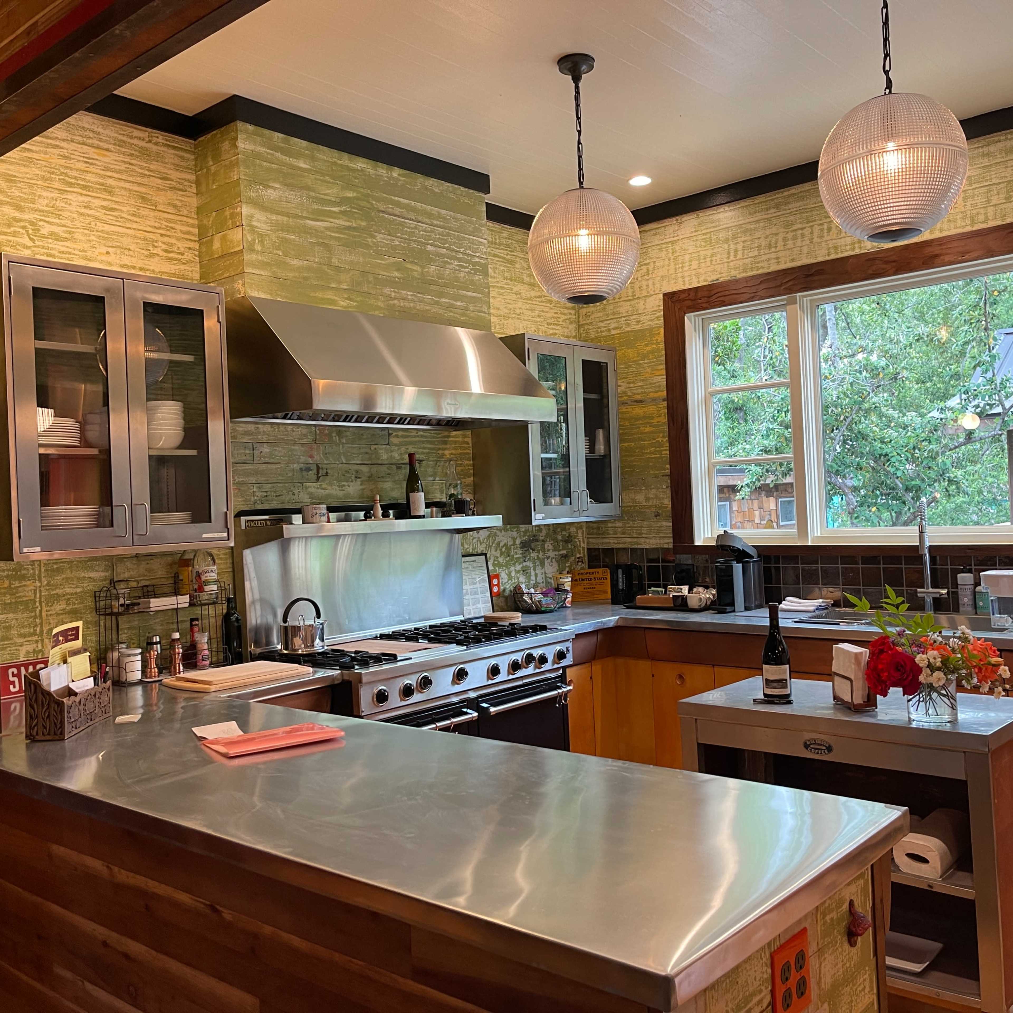 The kitchen features a stainless steel countertop, modern appliances, and green tiled walls, with a window overlooking a wooded area.