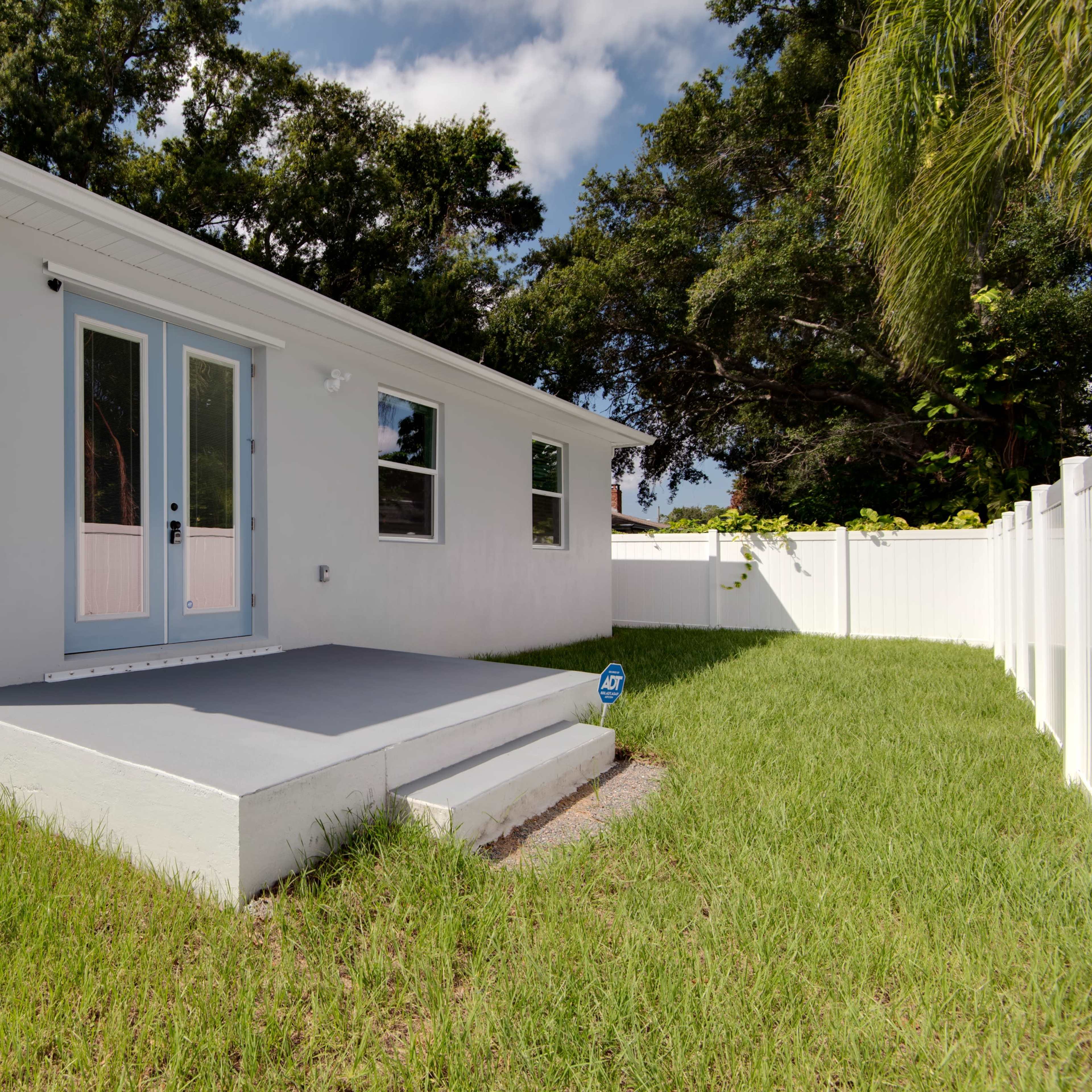 A modern single-story house with a white exterior, a small patio in the back, and a fenced yard with green grass.