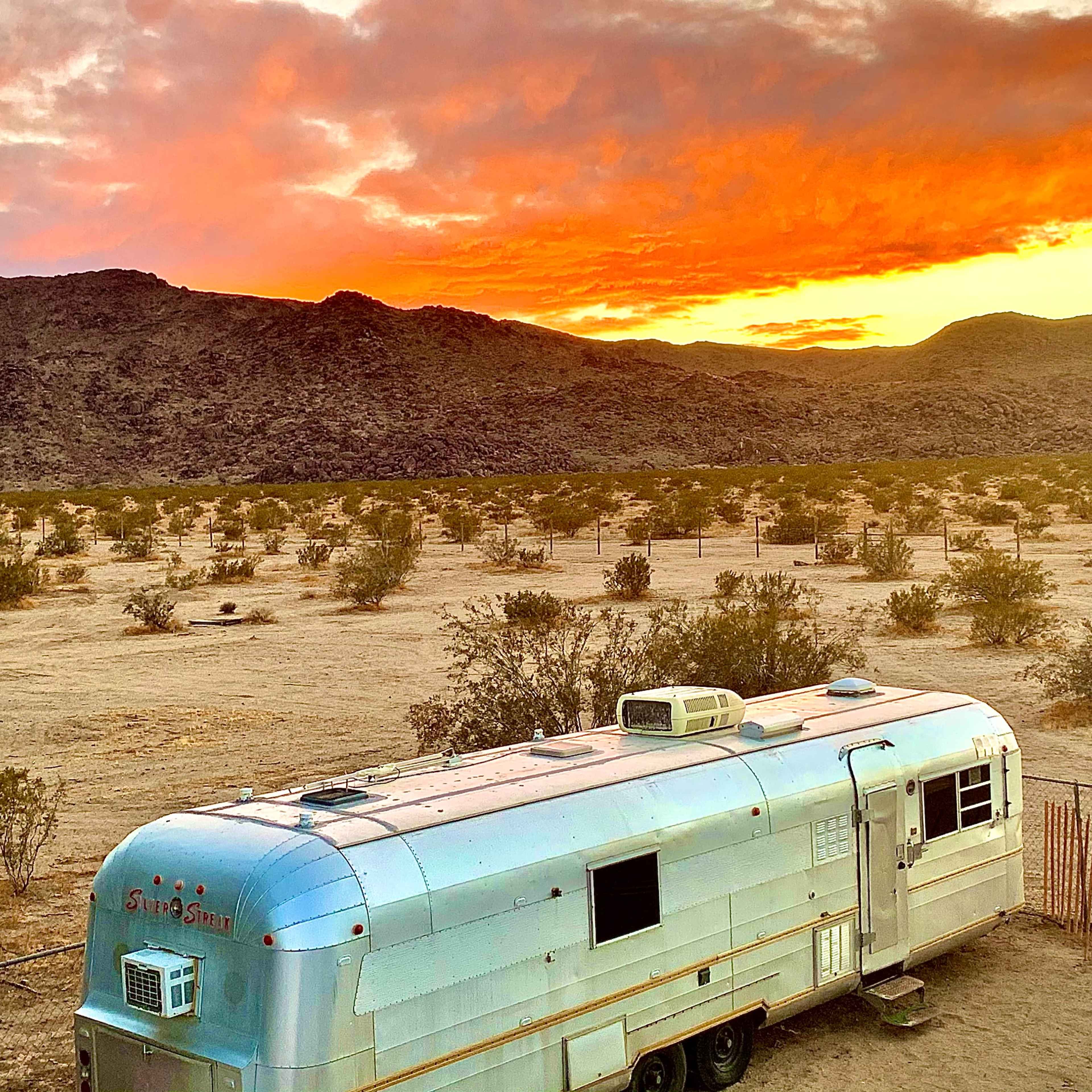 An Airstream trailer is parked in a desert landscape with a vibrant sunset illuminating the sky.