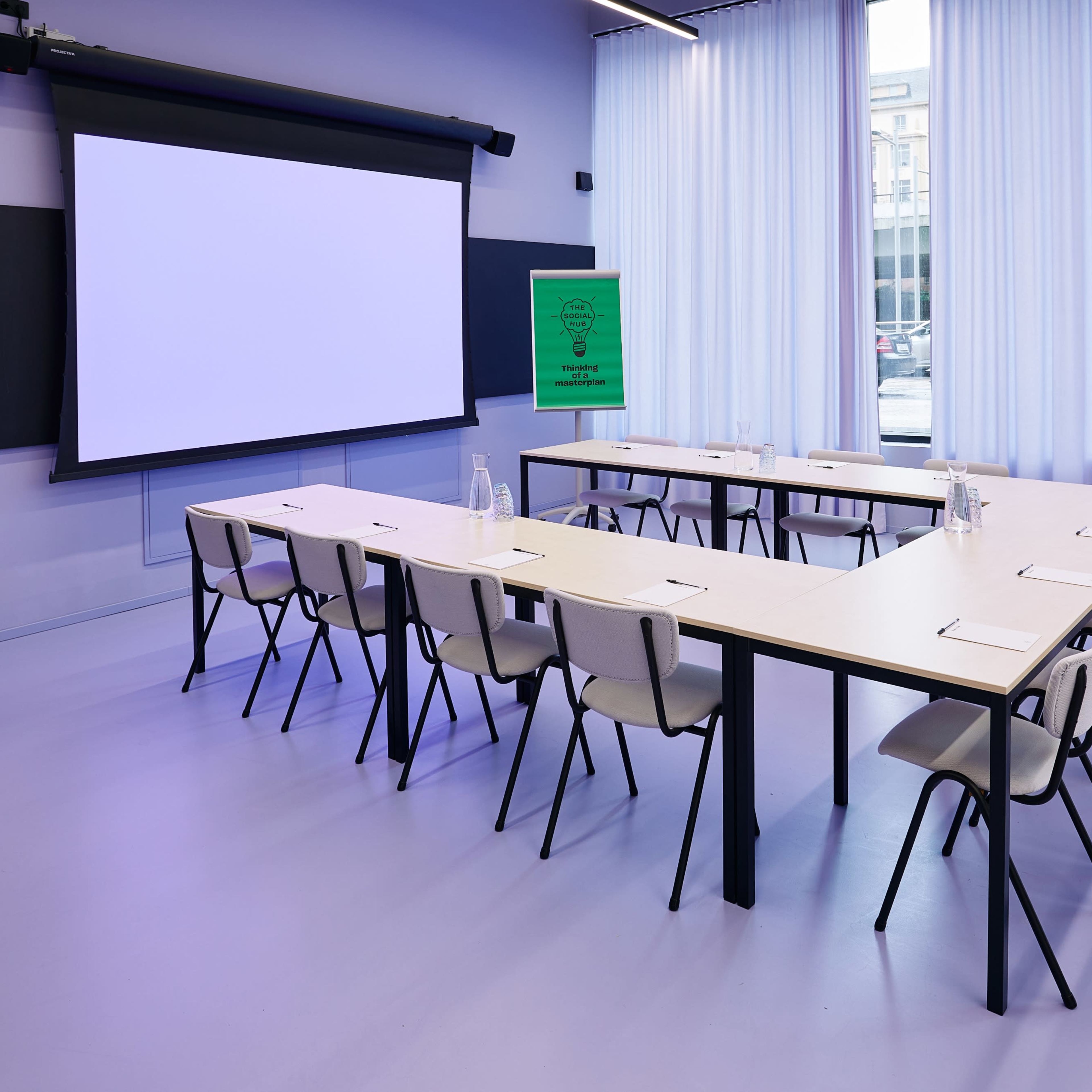 A modern conference room features a rectangular table surrounded by chairs, with a projector screen at the front and large windows allowing natural light.