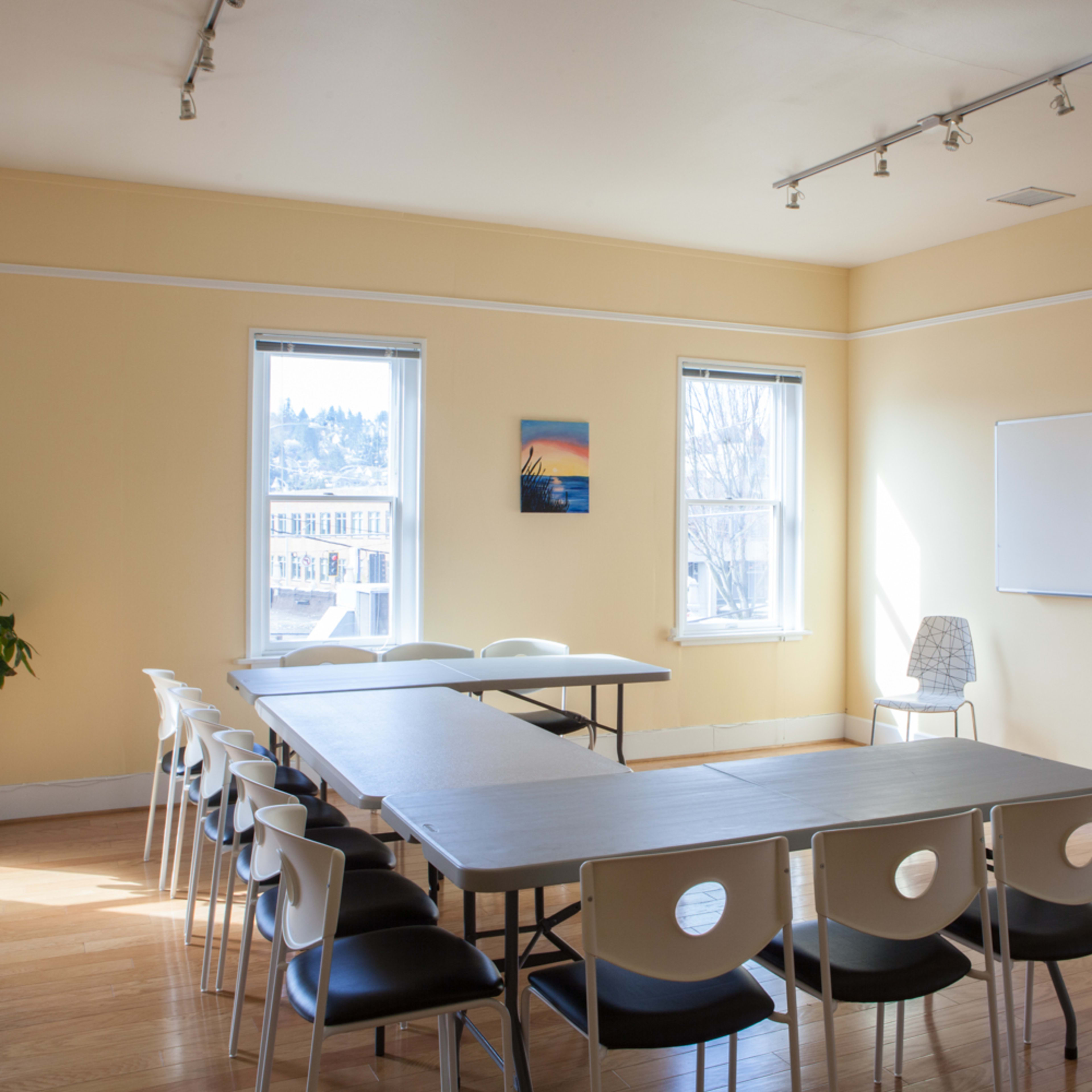 A well-lit meeting room features a long table surrounded by chairs, with windows providing natural light and a whiteboard on one wall.