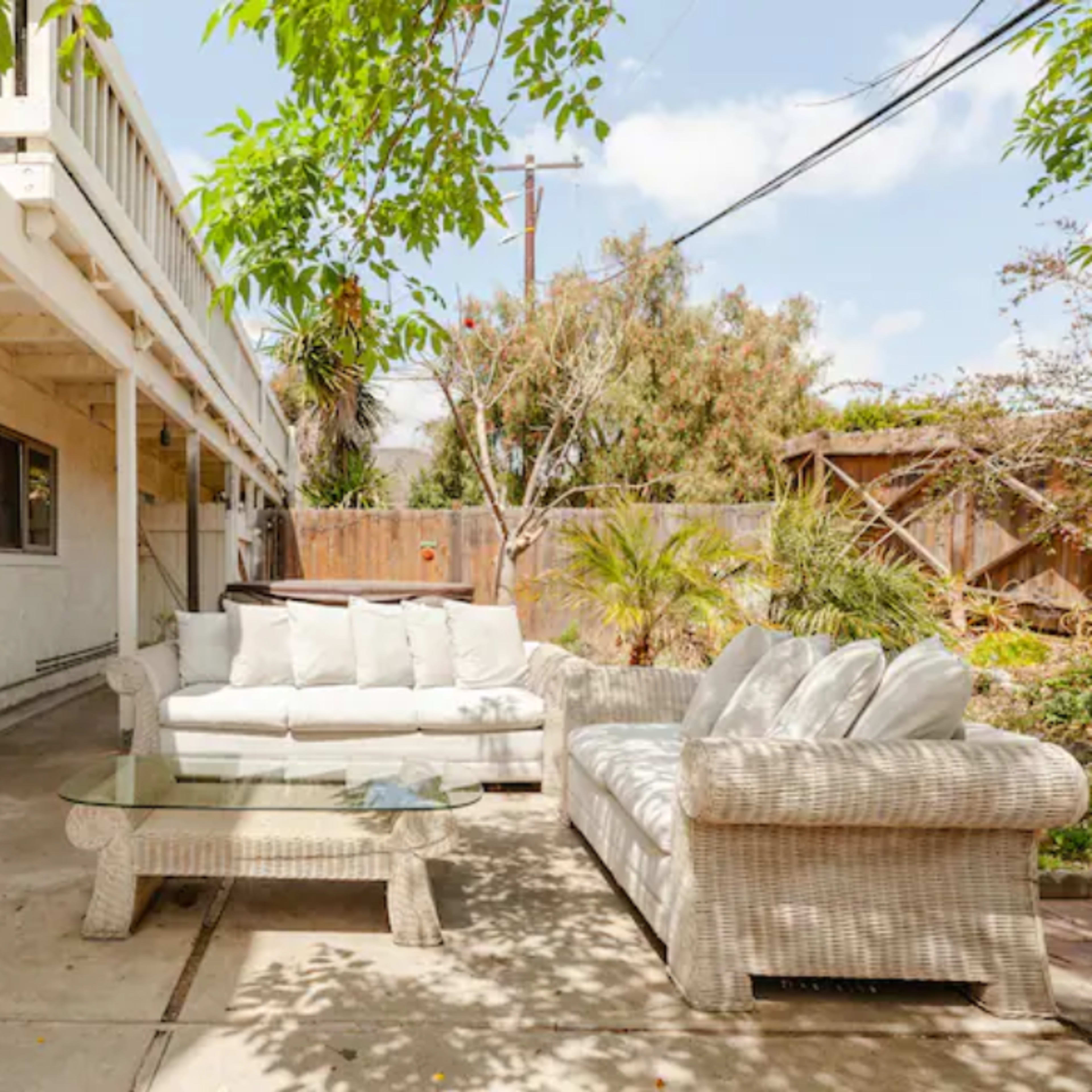 A patio area features a white wicker sofa set and a glass coffee table surrounded by greenery and a wooden fence.