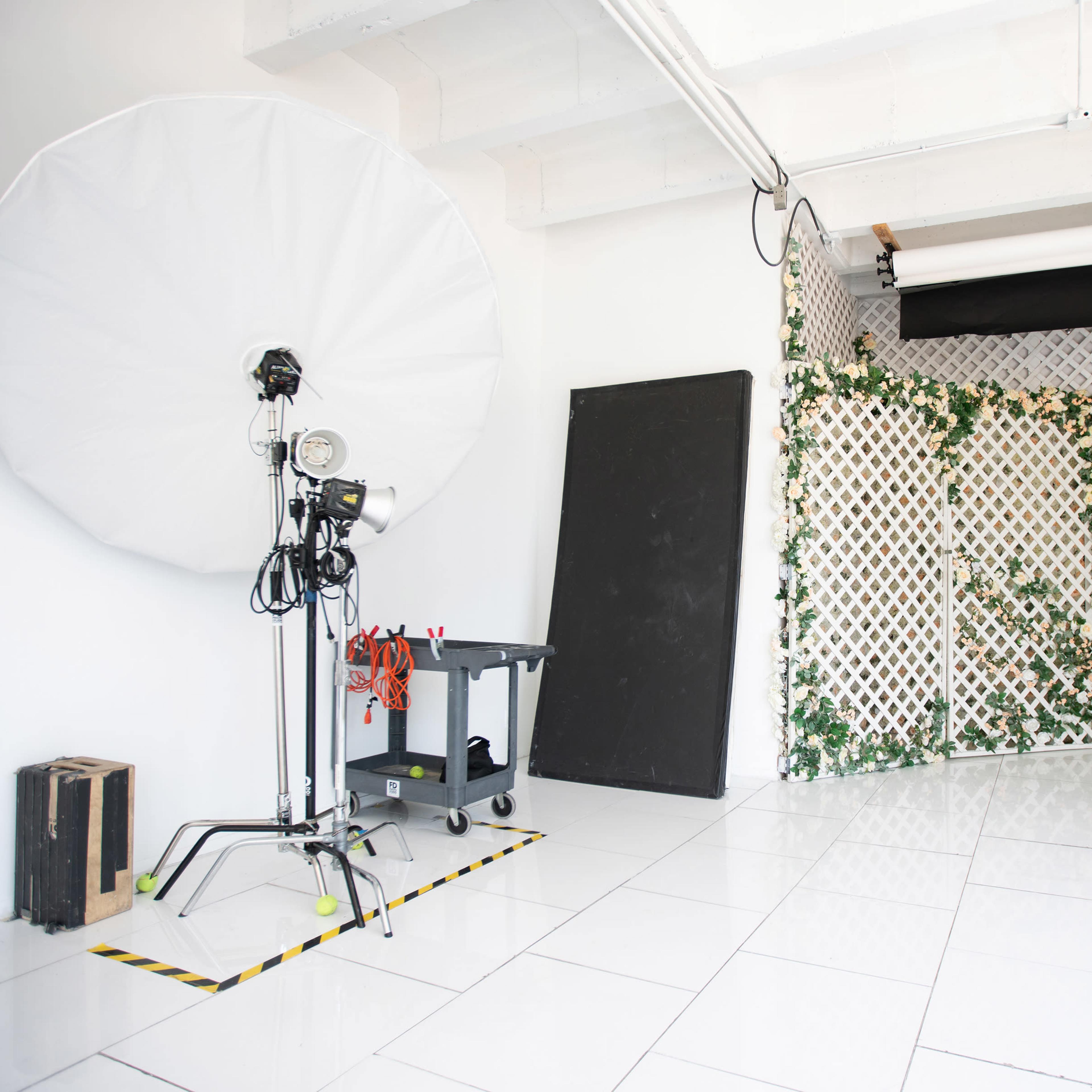 A photography studio features a large softbox, a backdrop with flowers, and a gray textured panel on white tiled flooring.