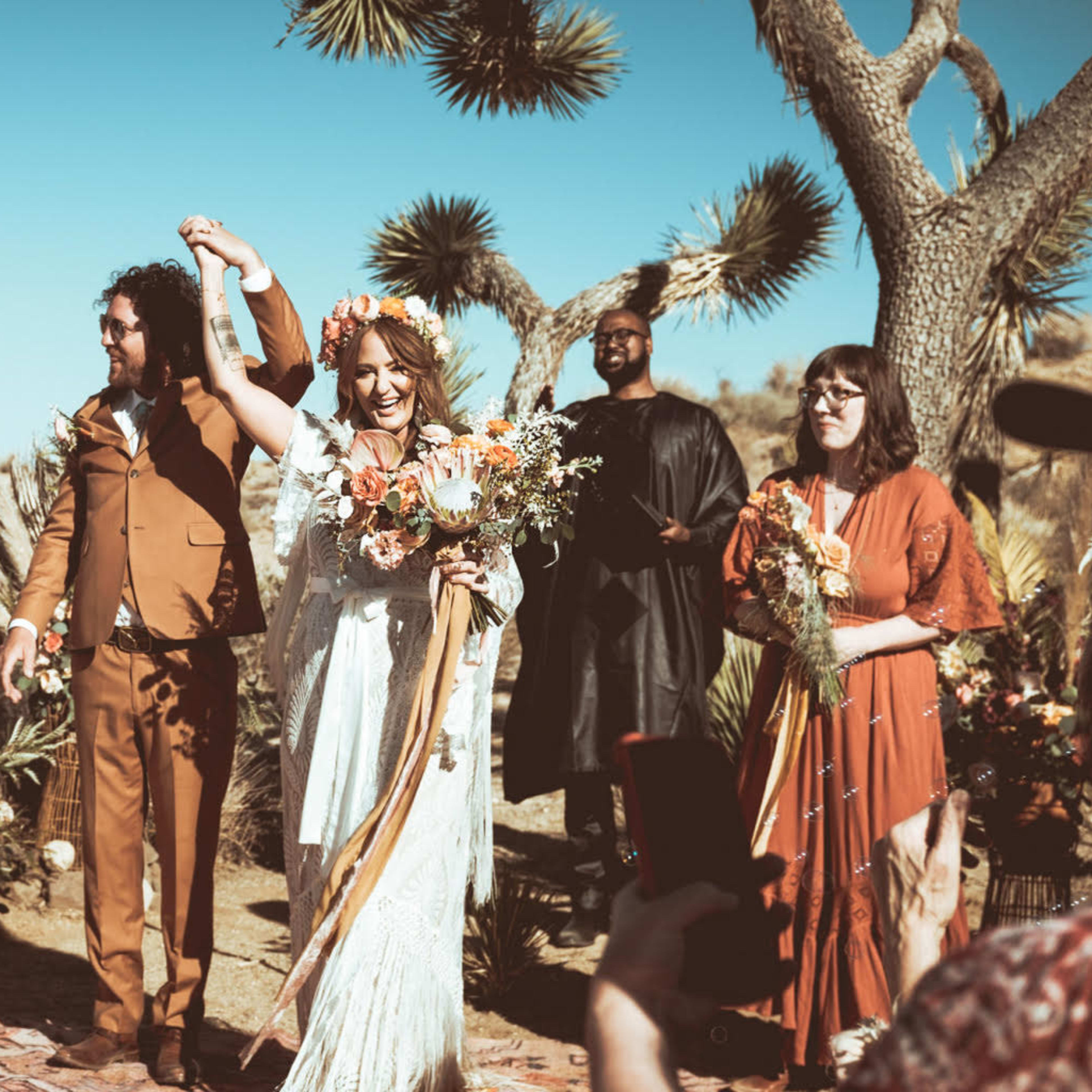 A couple celebrates their wedding under a clear blue sky in a desert setting, surrounded by guests holding flowers and drinks.