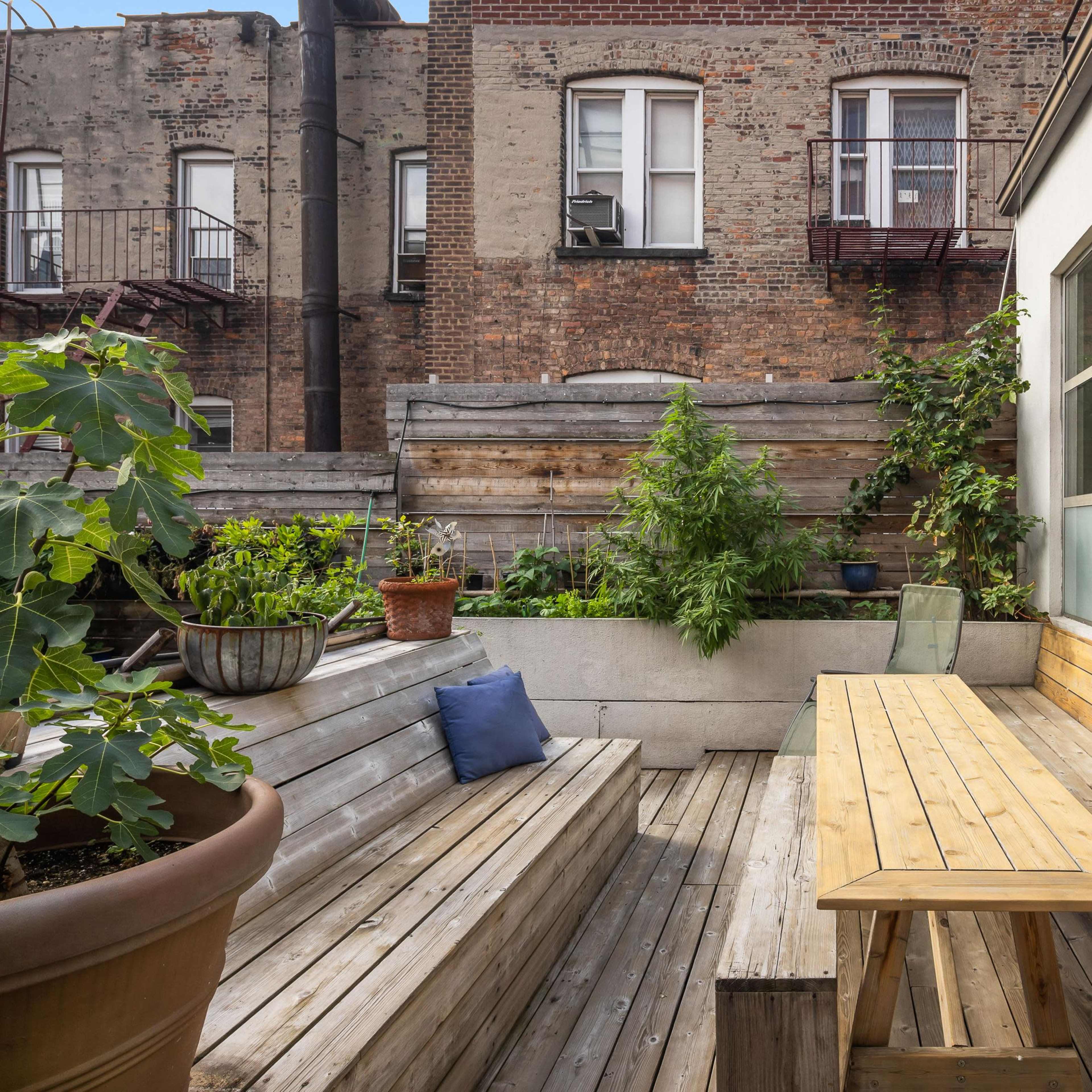 A wooden deck area with benches and a table, surrounded by potted plants, against a backdrop of brick buildings.