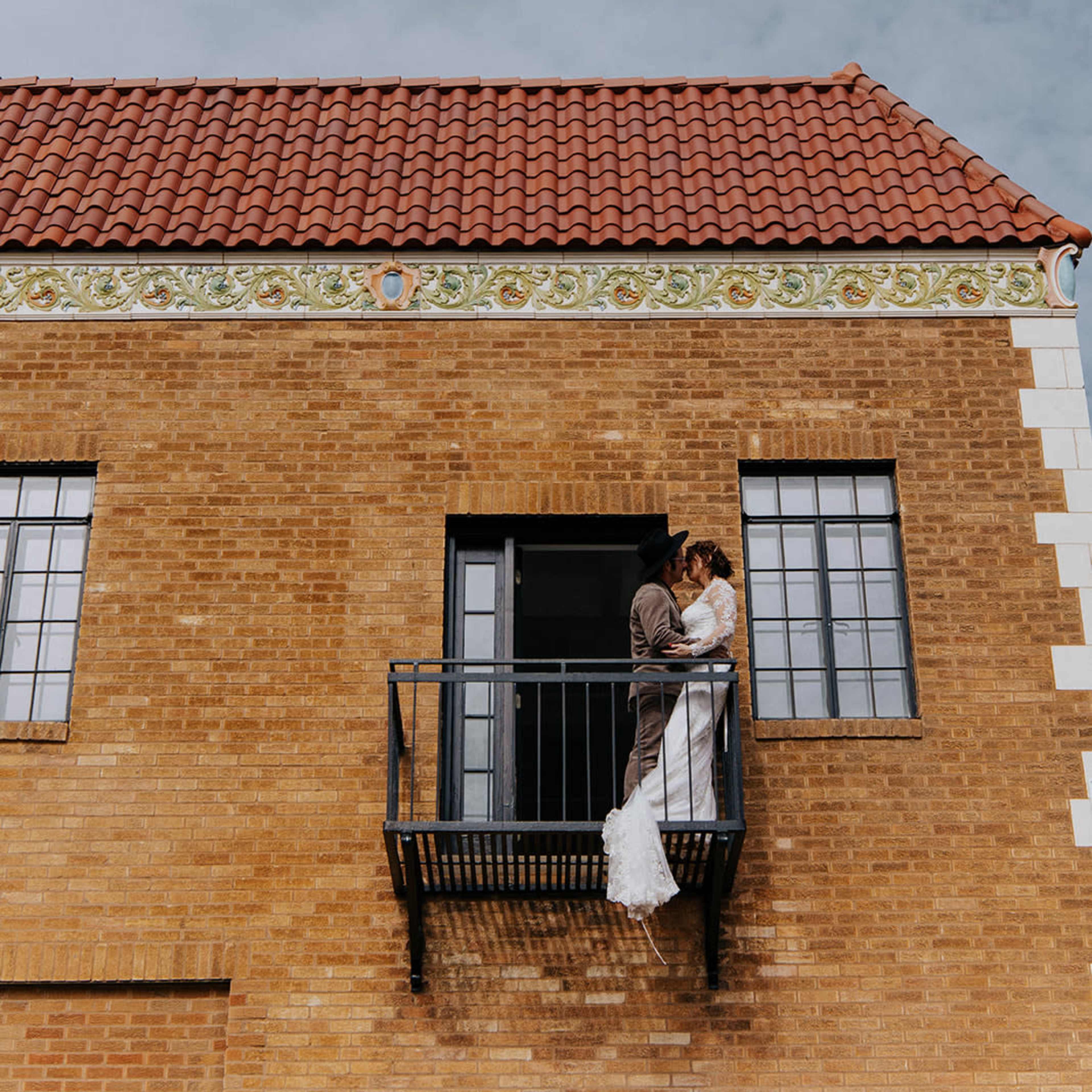 A couple in wedding attire embraces on a balcony of a brick building with a tiled roof.