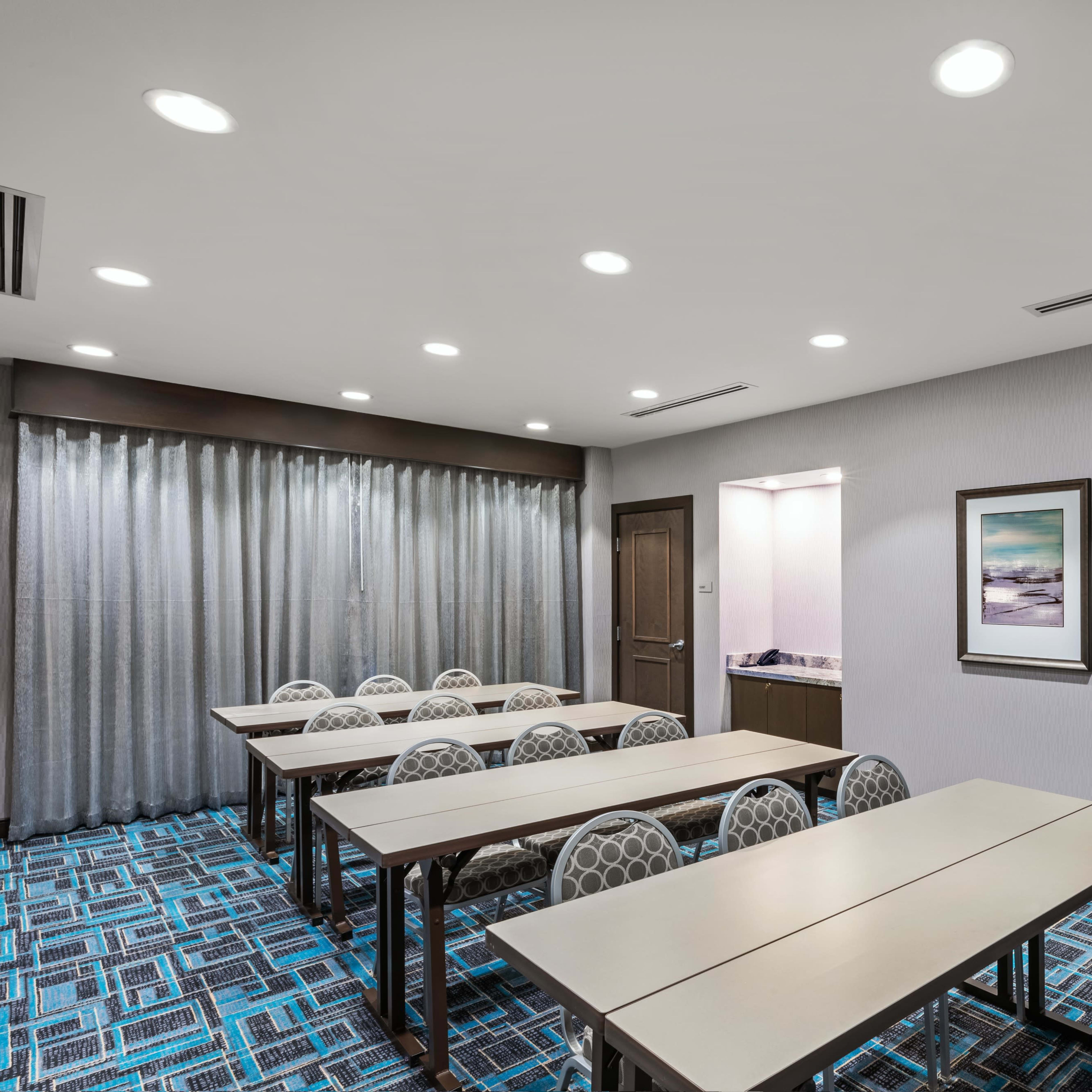A meeting room features several arranged tables with chairs around them, alongside large windows covered with curtains, and two framed artworks on the walls.