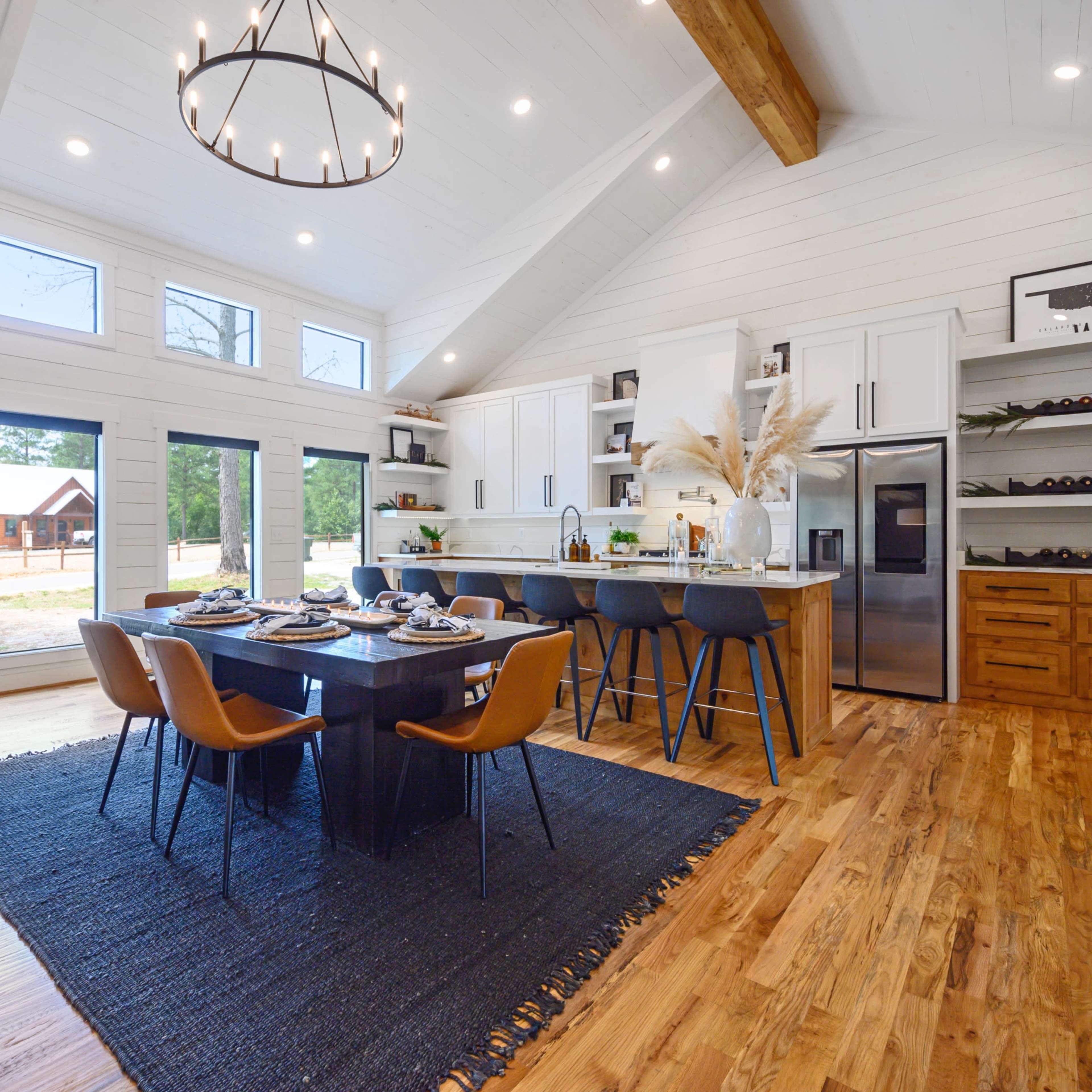 A modern open-concept kitchen and dining area features a wooden table surrounded by chairs, with large windows allowing natural light to fill the space.