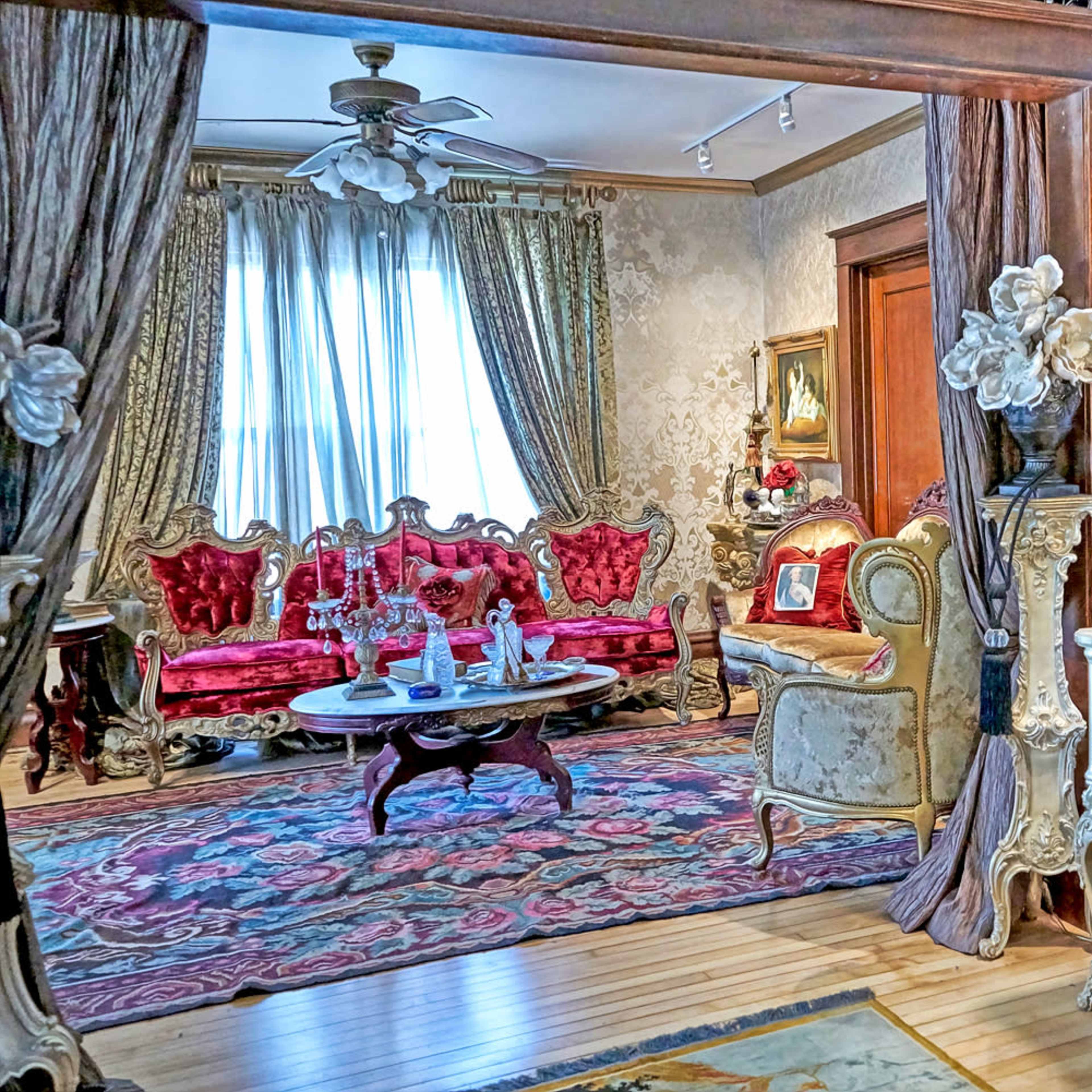 An ornate living room featuring plush red sofas, intricate decorations, a patterned rug, and a large window with draped curtains.