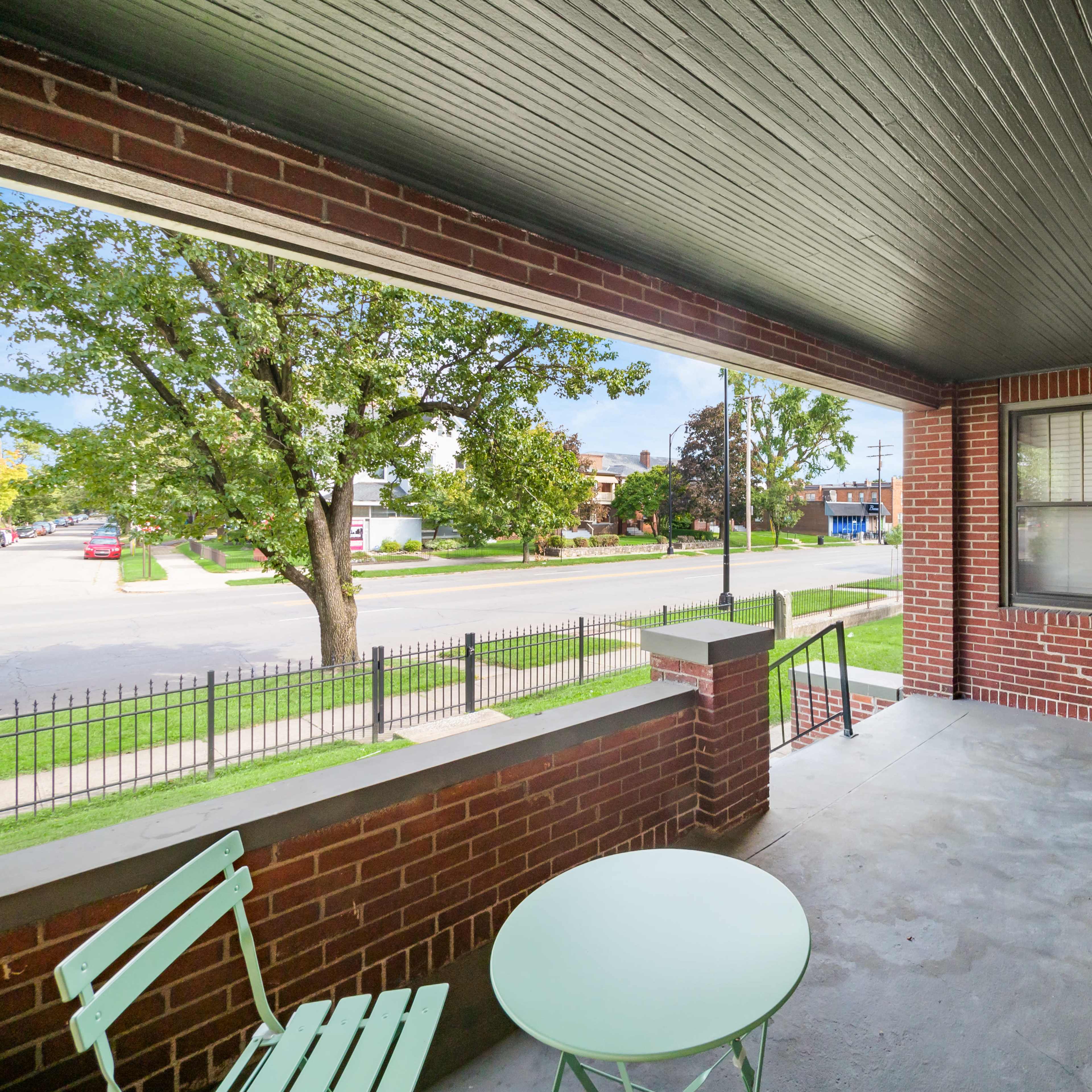 A covered porch with a green table and chairs, overlooking a tree-lined street.