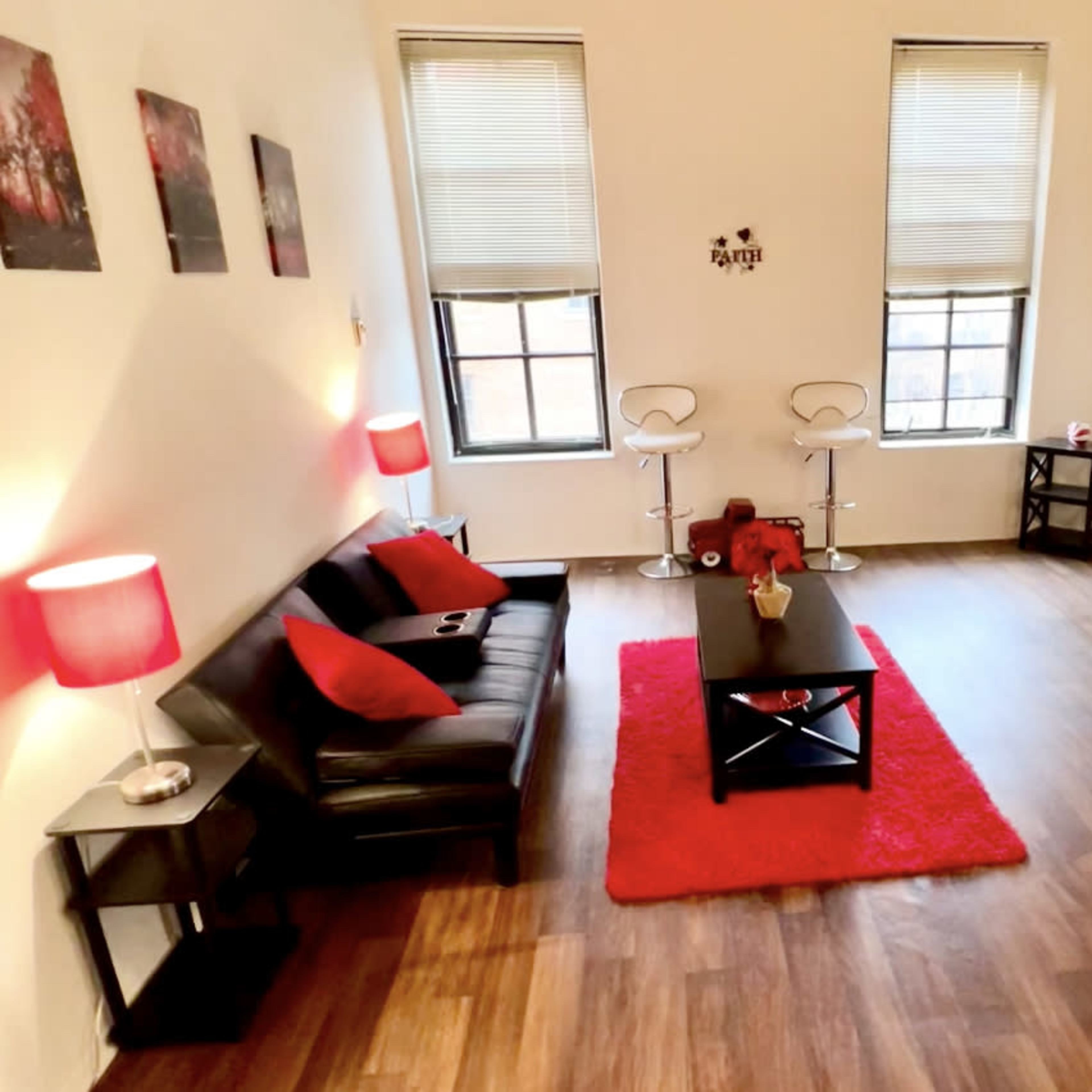 A minimalist living room with a black sofa, a red rug, two bar stools, and a coffee table, featuring large windows with blinds.