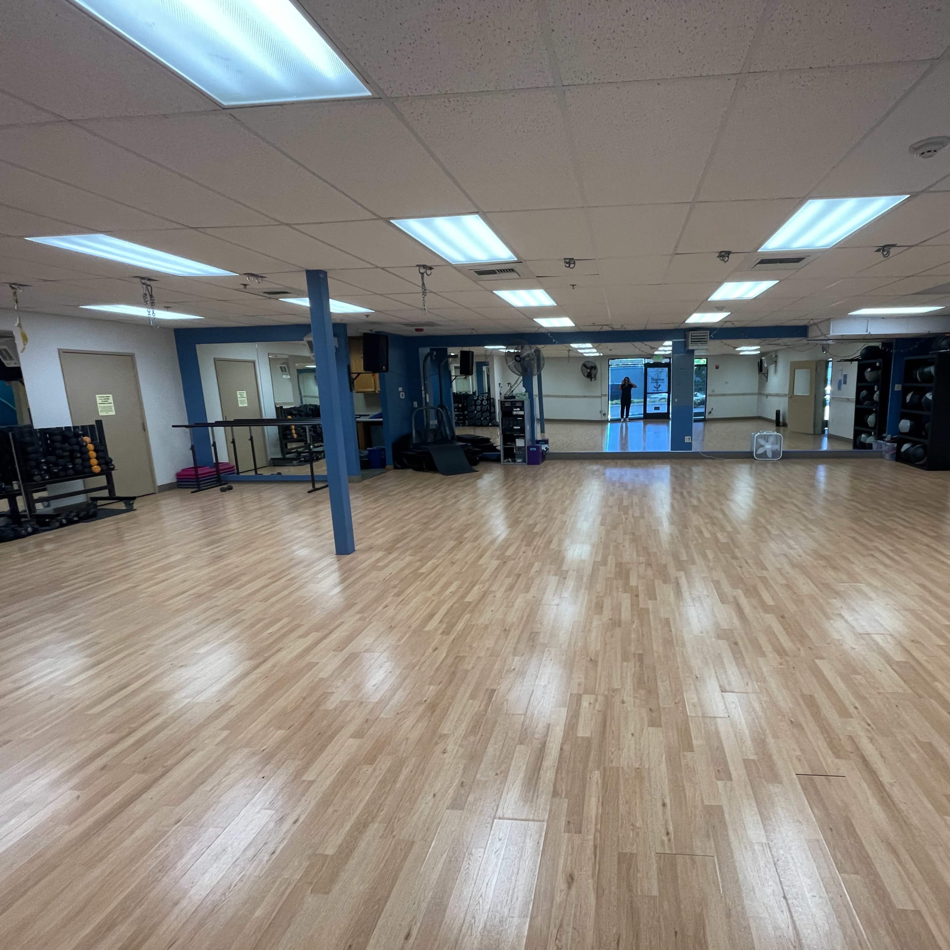 A spacious, clean exercise studio featuring wooden flooring, mirrors, and fitness equipment along the walls.